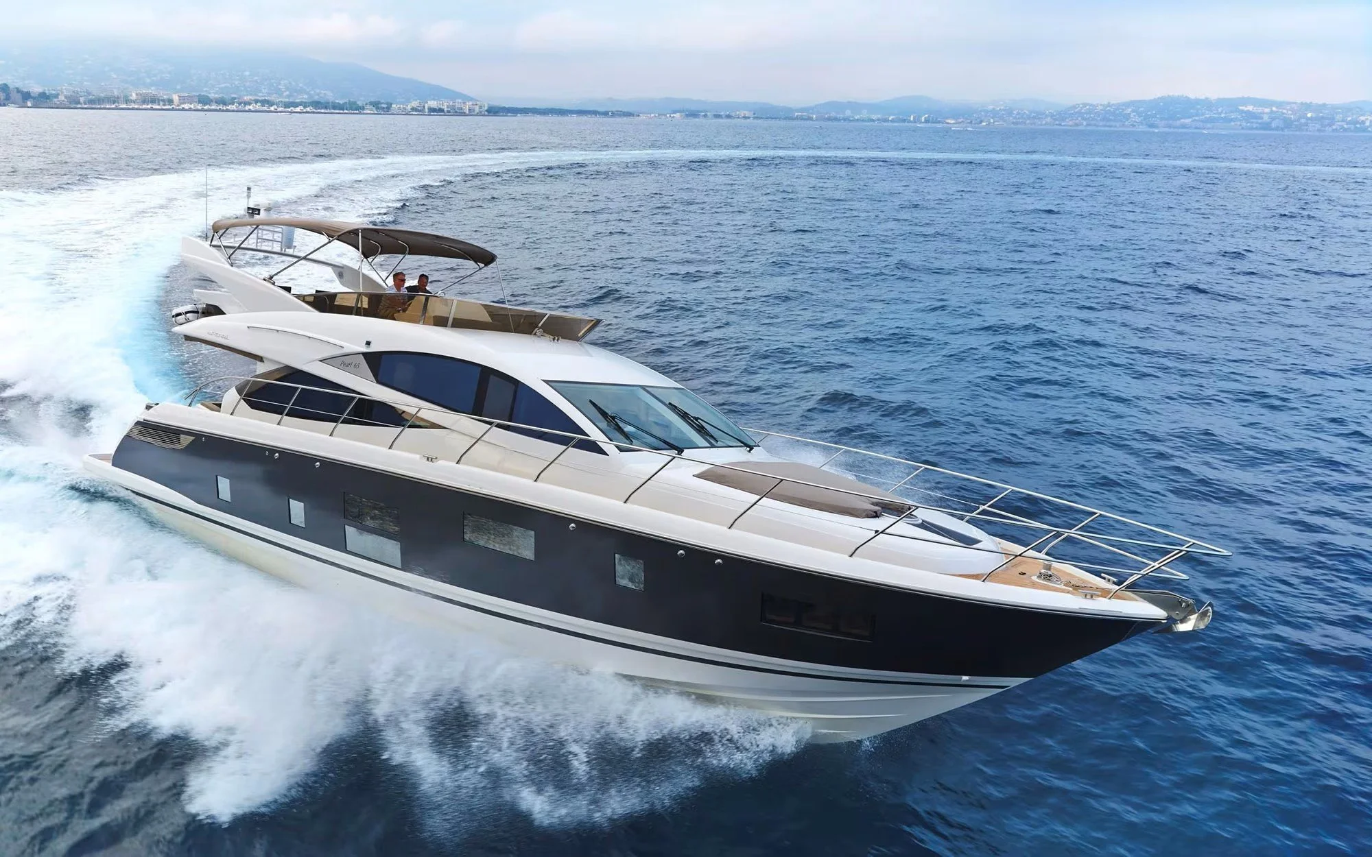 A luxury motor yacht cruising on the open water with two people on the upper deck, a cityscape and mountainous landscape in the background.