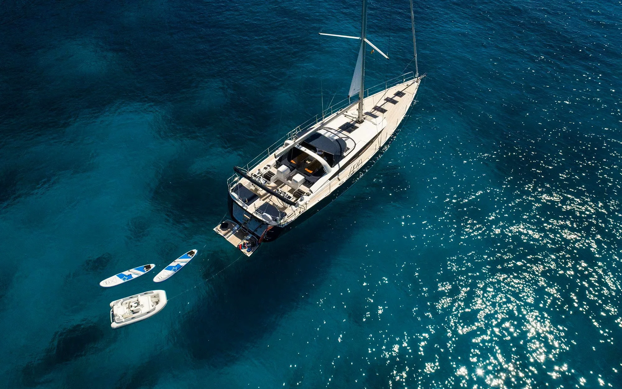 An aerial view of a sailboat anchored in the ocean, accompanied by a small inflatable boat and two paddleboards nearby.
