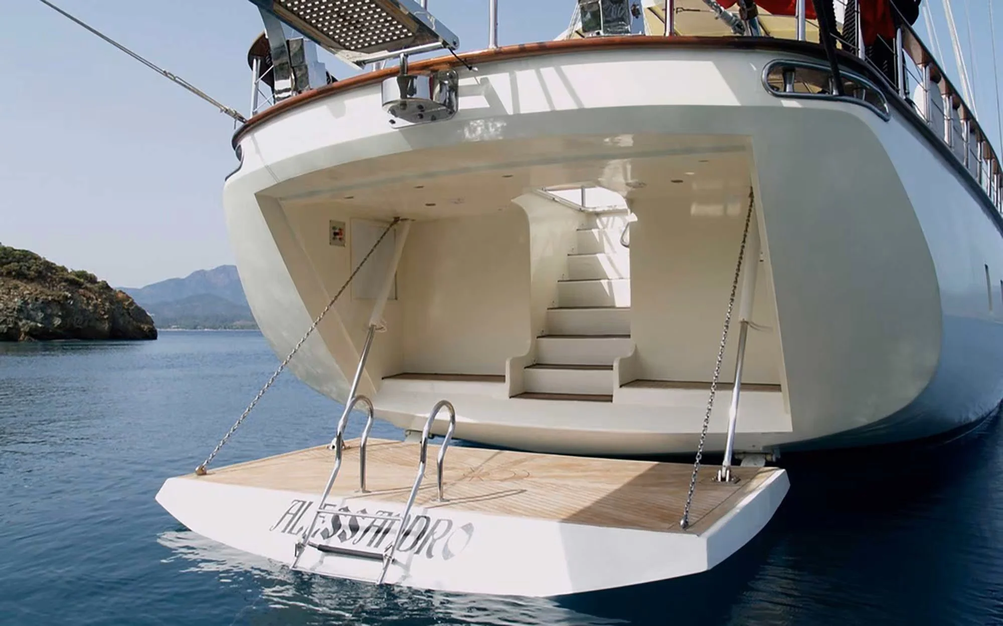 Back view of a luxury yacht with a wooden swim platform, stairs leading to the deck, and a calm body of water and distant mountains in the background.
