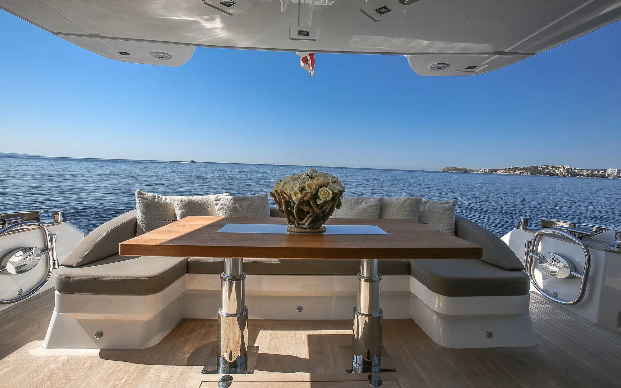 Interior of a yacht with a table decorated with a flower arrangement, overlooking open water and a distant shoreline under a clear blue sky.