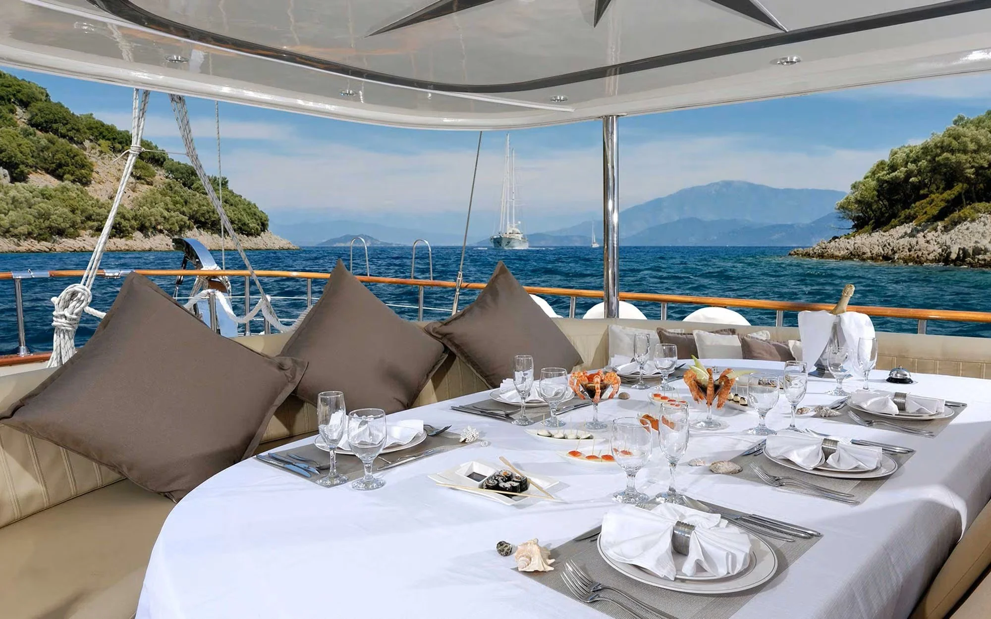 Elegant dining table set for a meal on a yacht deck, with white tablecloth, napkins, glassware, and shells in the foreground, with a view of the sea, mountains, and sailboats in the distance.