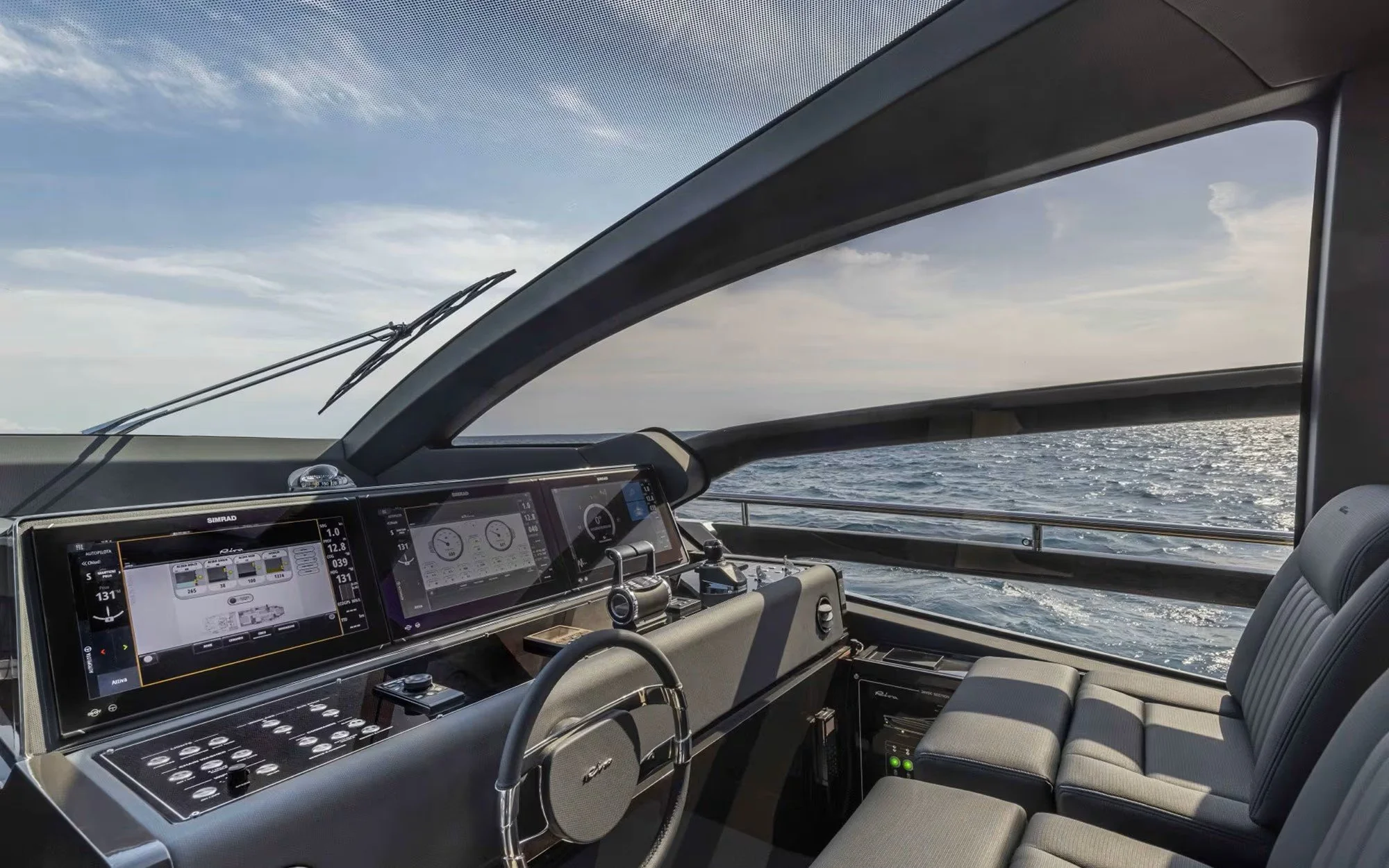 Interior of a luxury yacht with a dashboard of modern digital navigation and control screens, a steering wheel, and a seating area by the window overlooking the open sea.