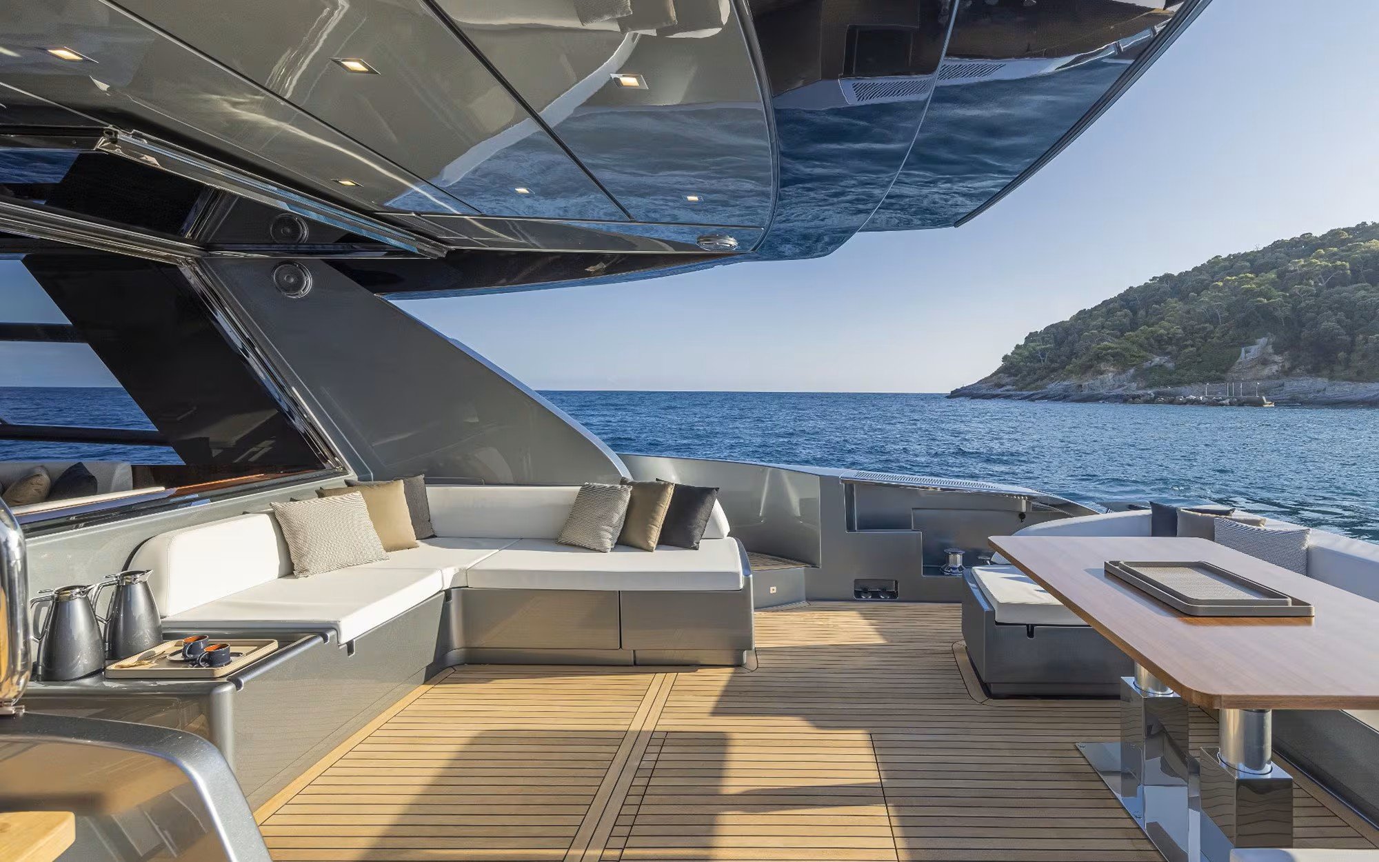Outdoor deck area of a yacht with sea and a hillside in the background, featuring a white cushioned seating area, a wooden table, and some accessories.