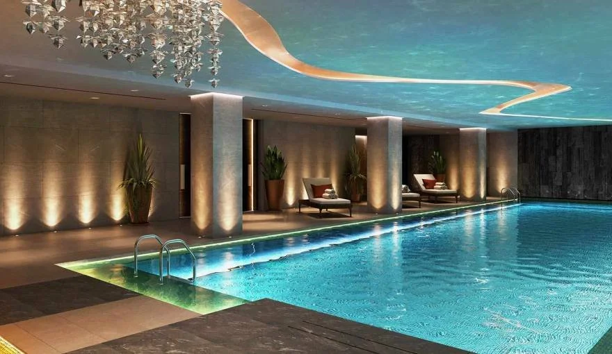 Indoor swimming pool with ambient lighting, lounge chairs, and potted plants under a decorative ceiling.