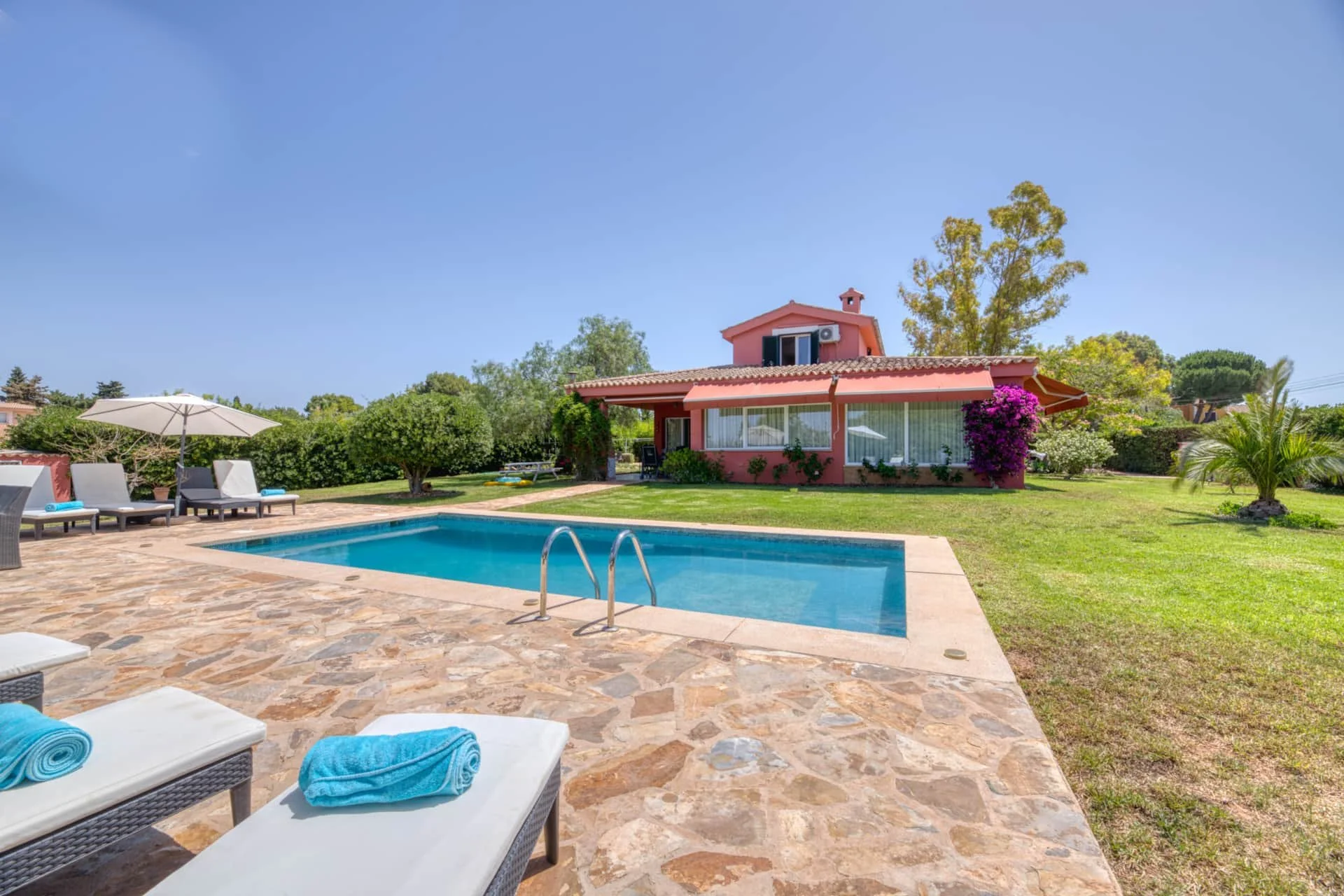 A backyard with a swimming pool, lounge chairs with towels, a large umbrella, a grassy area, trees, bushes, and a pink two-story house with a tiled roof and an enclosed porch.