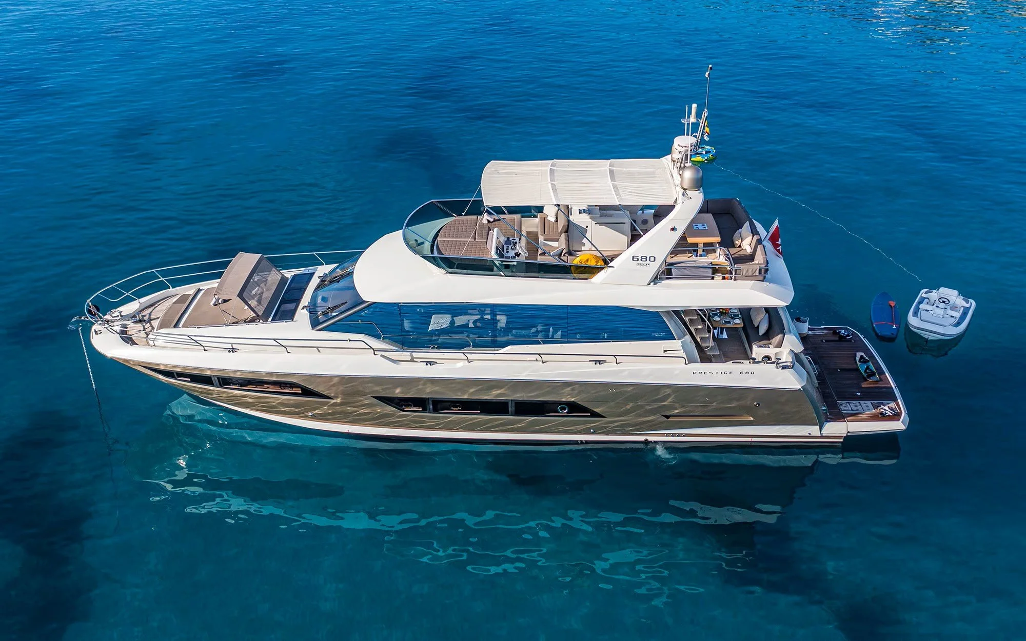Aerial view of a luxury yacht with a small boat and paddleboard nearby, floating on calm blue water.
