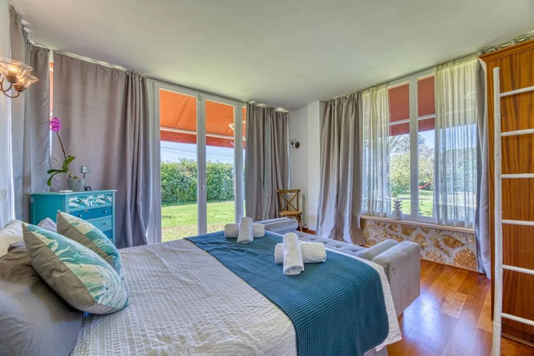 Bright bedroom with large windows and sliding glass door overlooking a green lawn. The bed has white and blue bedding with rolled towels. There's a turquoise dresser, a wooden chair, and a wooden wardrobe. Curtains and sheer drapes adorn the windows.