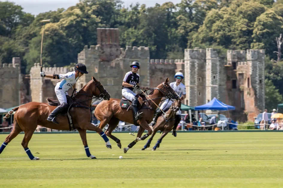 Three polo players riding horses and hitting a ball during a match on a field with a castle in the background.