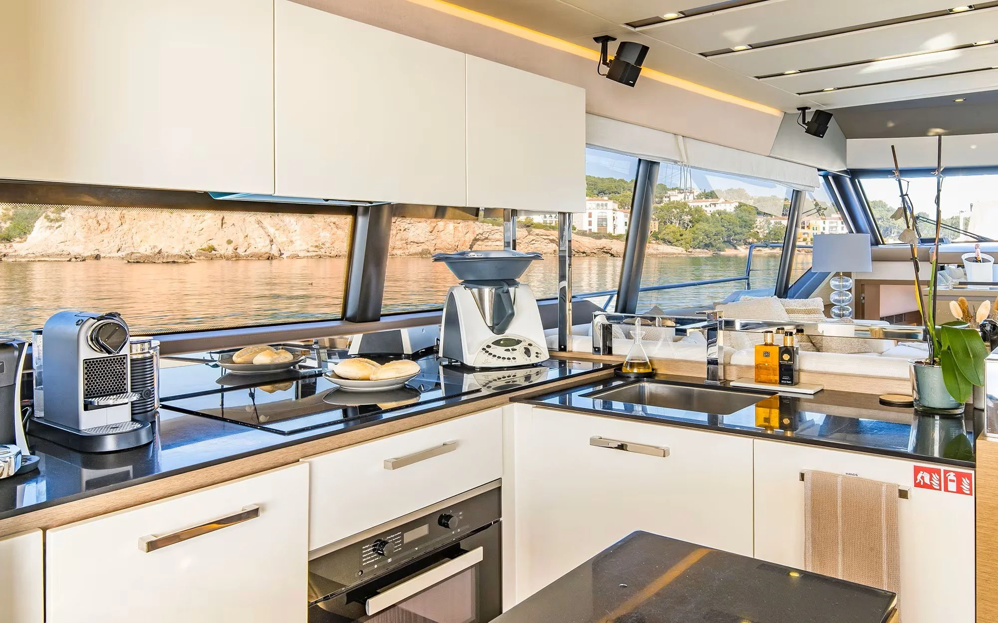 Modern yacht kitchen with a view of the water and shoreline, featuring white cabinets, black countertops, and various kitchen appliances.
