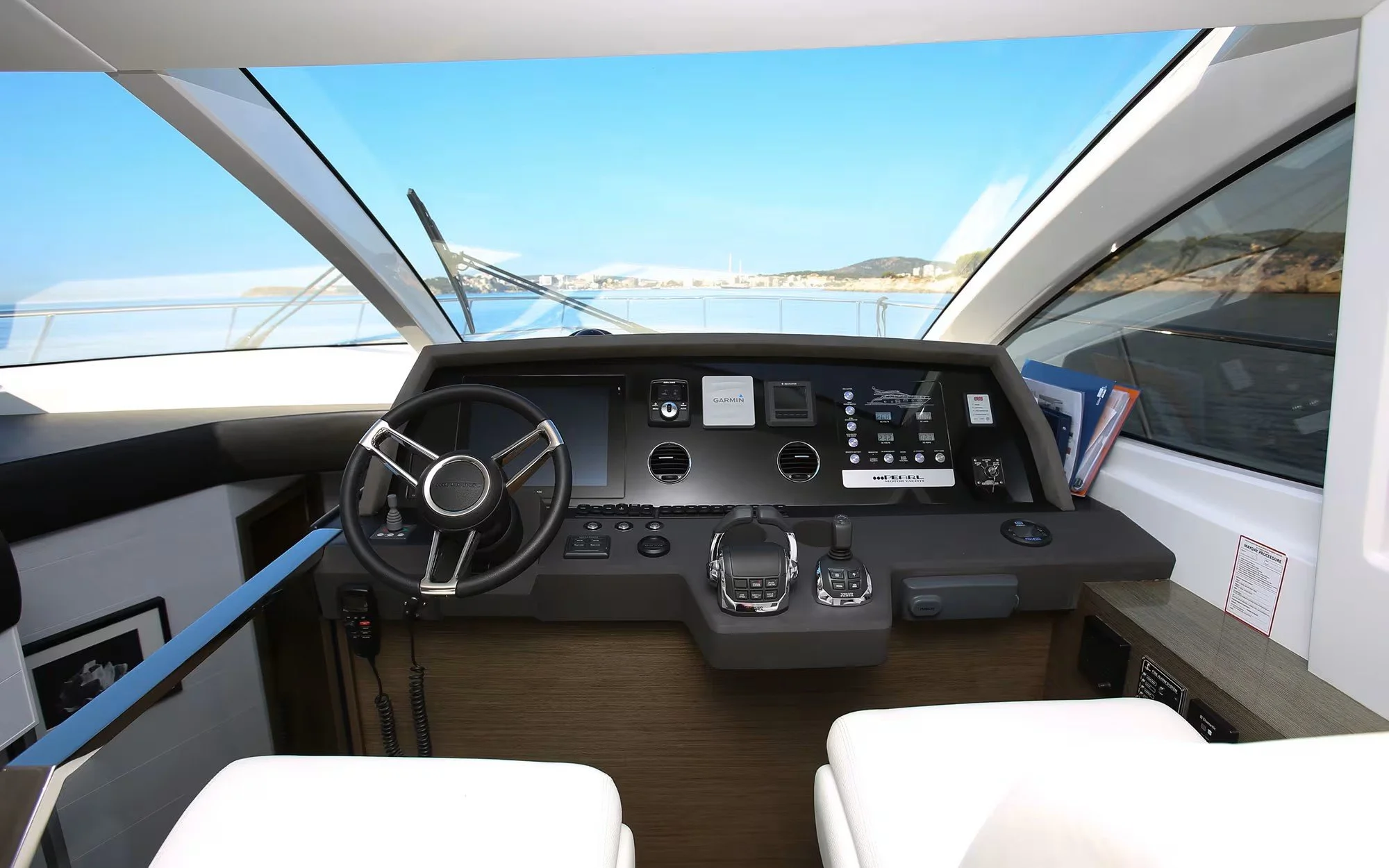 View of a yacht's helm with steering wheel, navigation screens, controls, and a view of the ocean and coastline through large windows