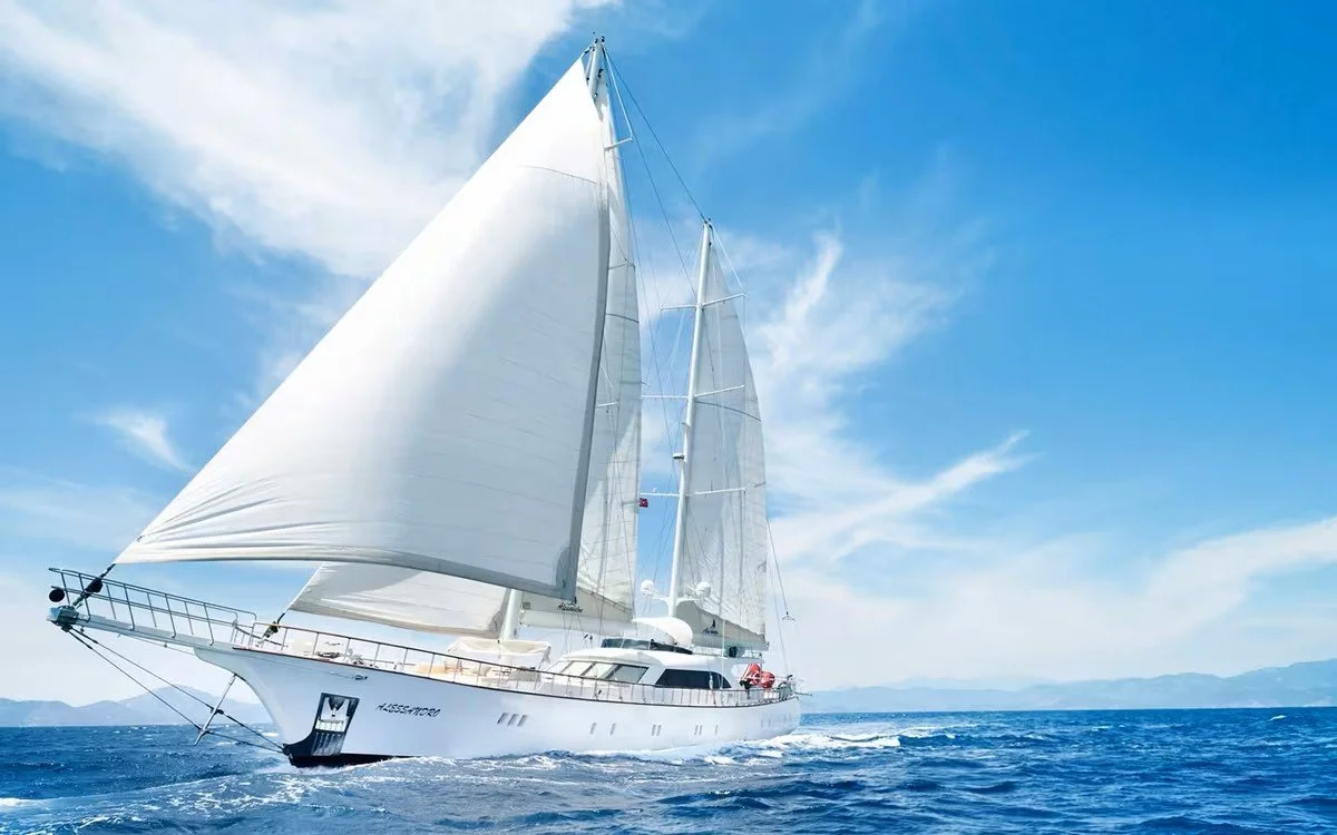 White sailboat with multiple sails on open water under a blue sky with scattered clouds.