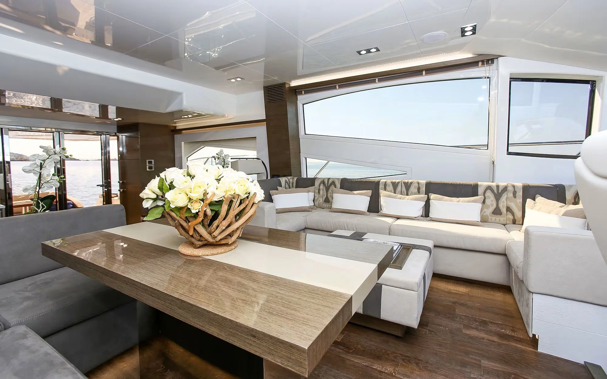 Interior of a yacht lounge with a large, curved sofa, a wooden table with a floral arrangement, and expansive windows overlooking water.