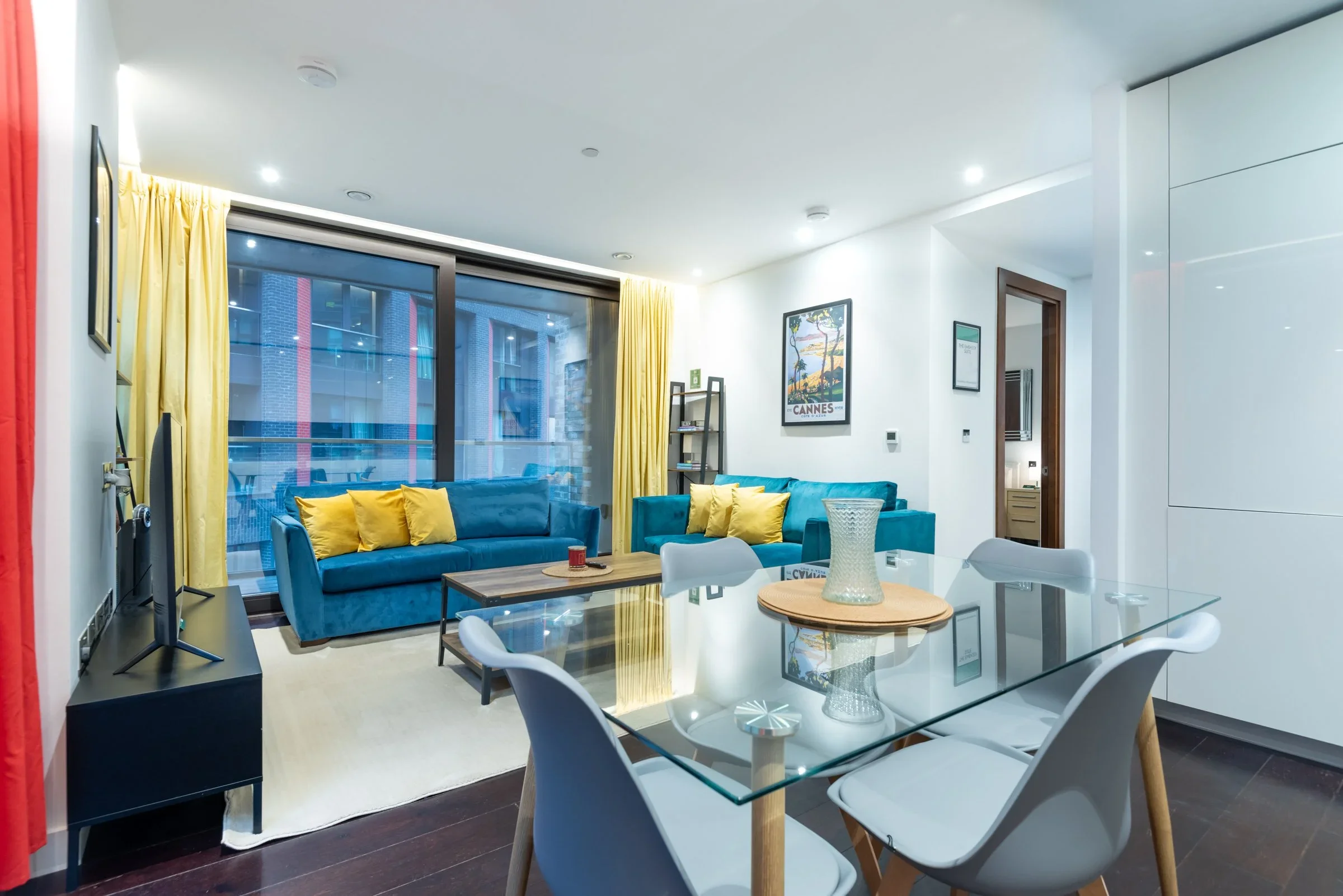 Modern living room with yellow curtains, blue sofas with yellow pillows, a glass dining table with white chairs, a wall-mounted TV, a rug, and framed posters on the wall.