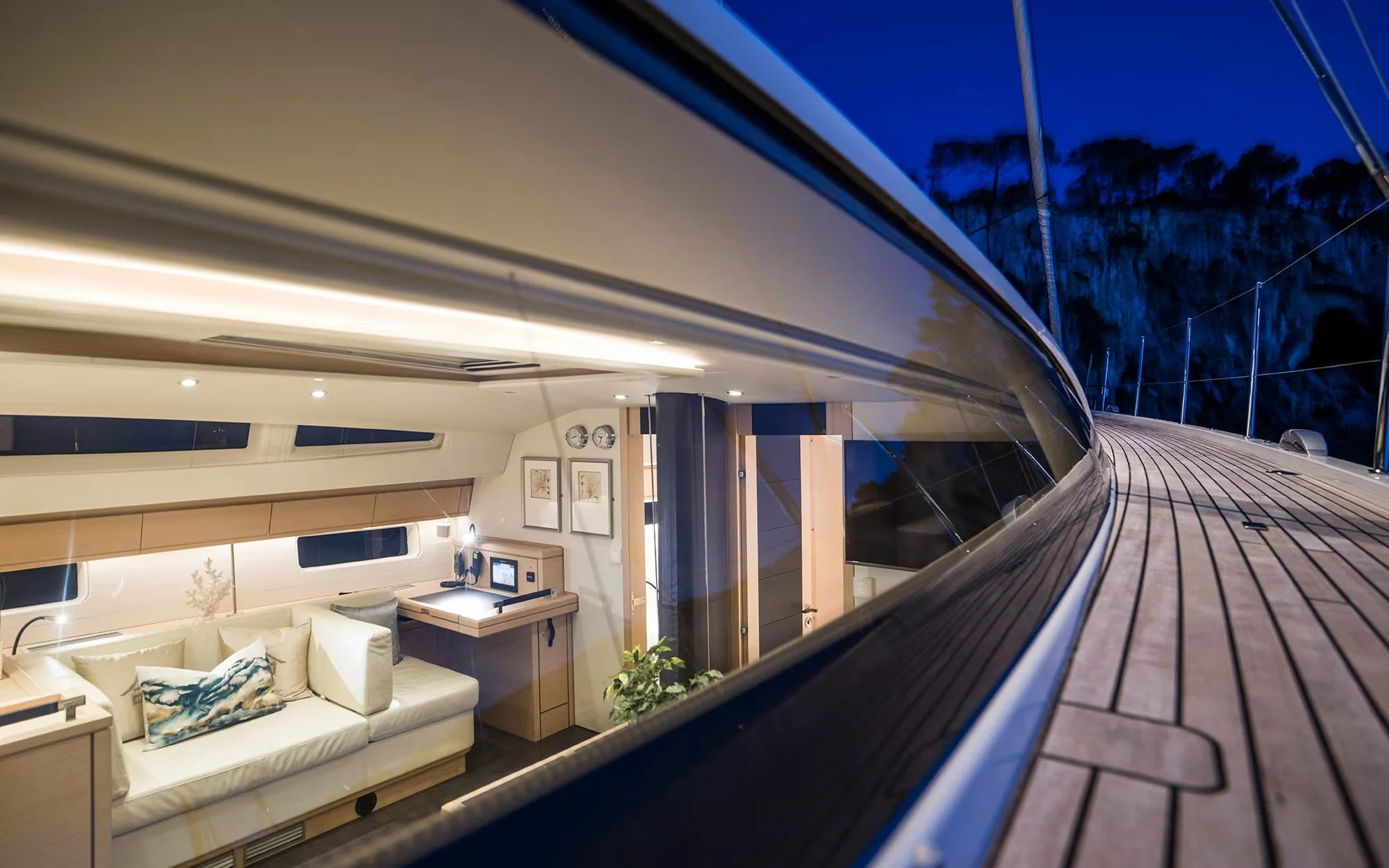 Interior view of a luxury yacht with a white sofa, wooden cabinets, and a computer, seen through the yacht's window at dusk or night.