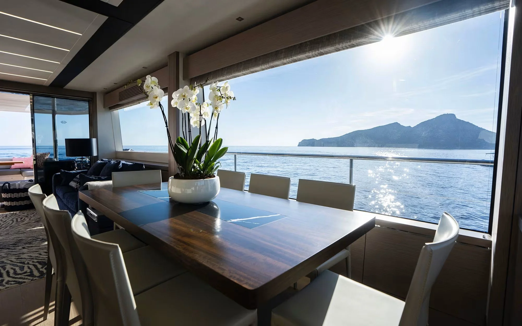 Interior of a modern yacht with a dining area overlooking the ocean and an island in the distance.