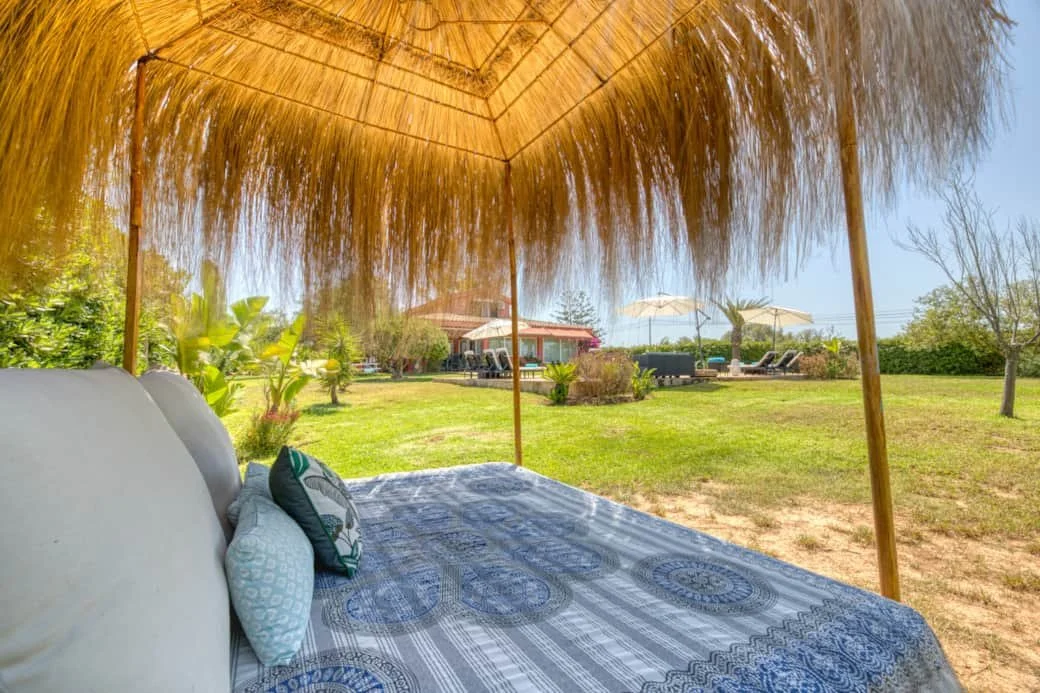 Outdoor bed with pillows under a thatched roof, overlooking a grassy yard with lounge chairs and umbrellas, and a house in the background.