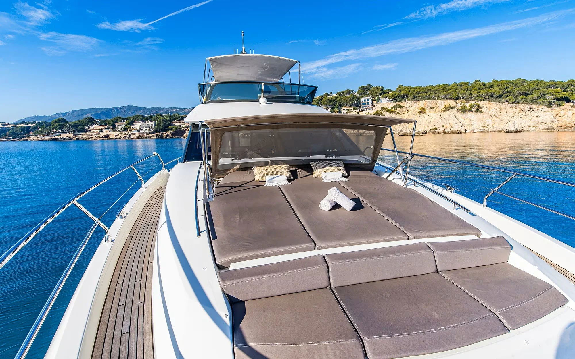 View of a luxury yacht anchored near a rugged shoreline under a clear blue sky, with a deck featuring sunbathing cushions and rolled towels.