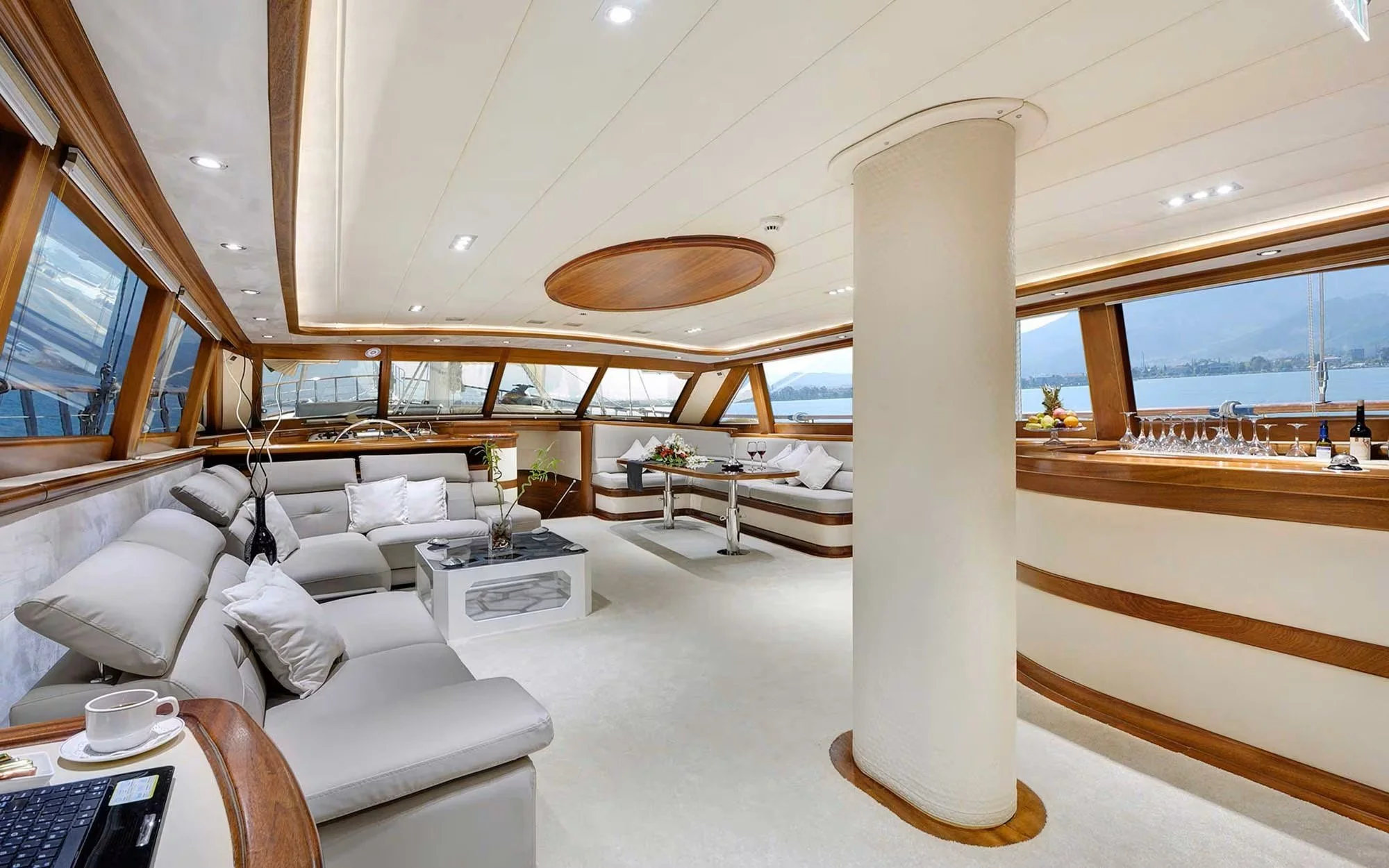 Luxurious yacht interior with large windows, white leather seating, a bar area with glasses and bottles, and a table with a flower arrangement.