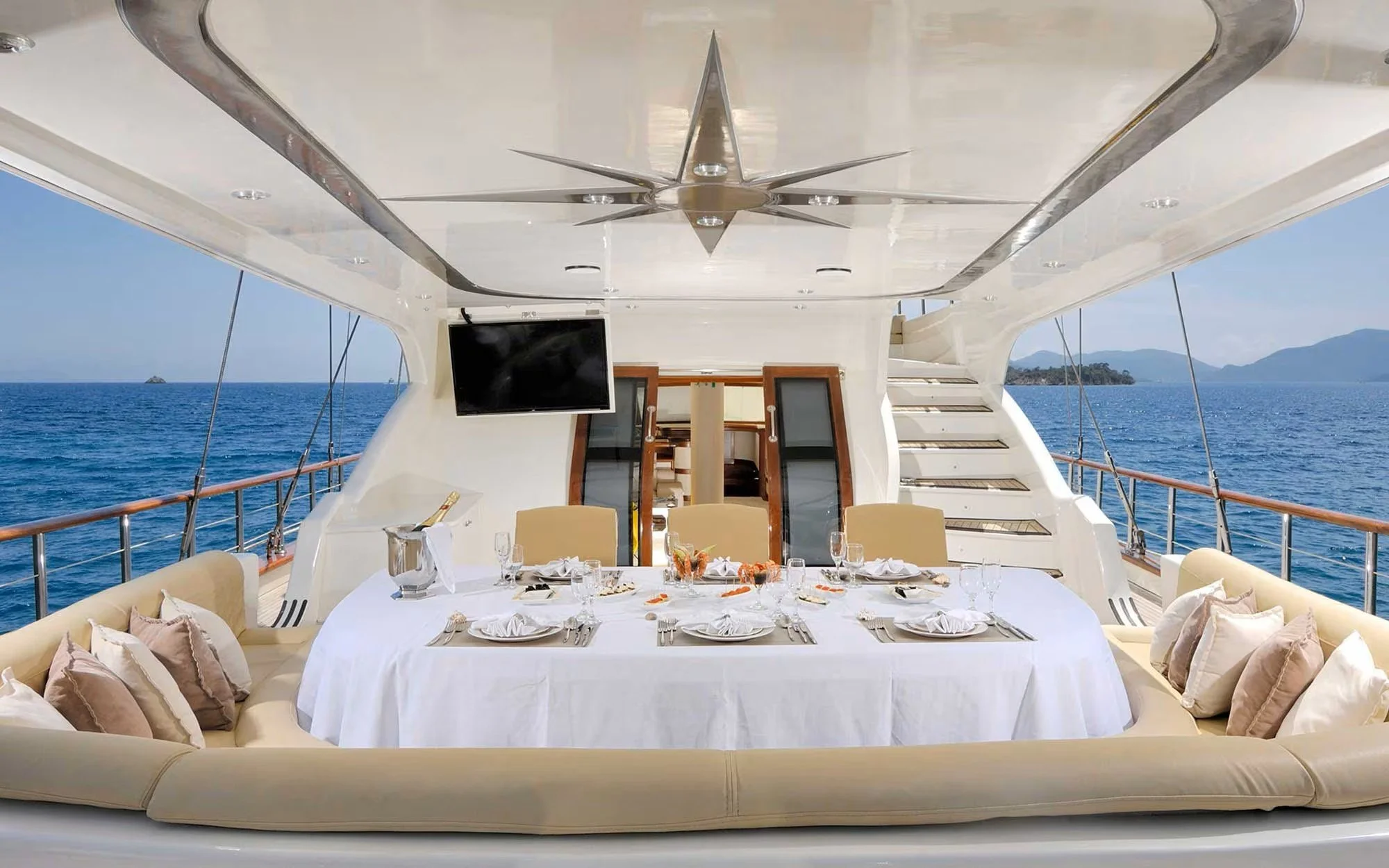 Dining area on a yacht with a table set with plates, glasses, and napkins, overlooking the ocean with islands in the distance.