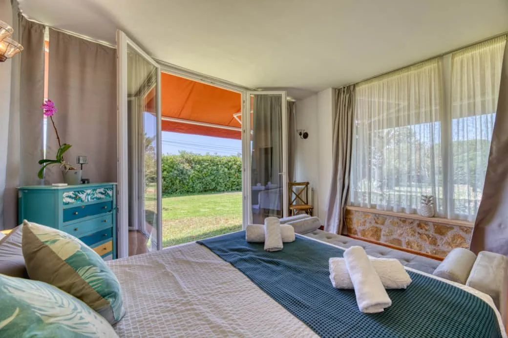 A cozy bedroom with a bed, pillows, and neatly folded towels. The room has large windows with sheer curtains, allowing natural light and a view of a green yard outside. There's a small teal dresser on the left and a wooden chair near the corner. An o