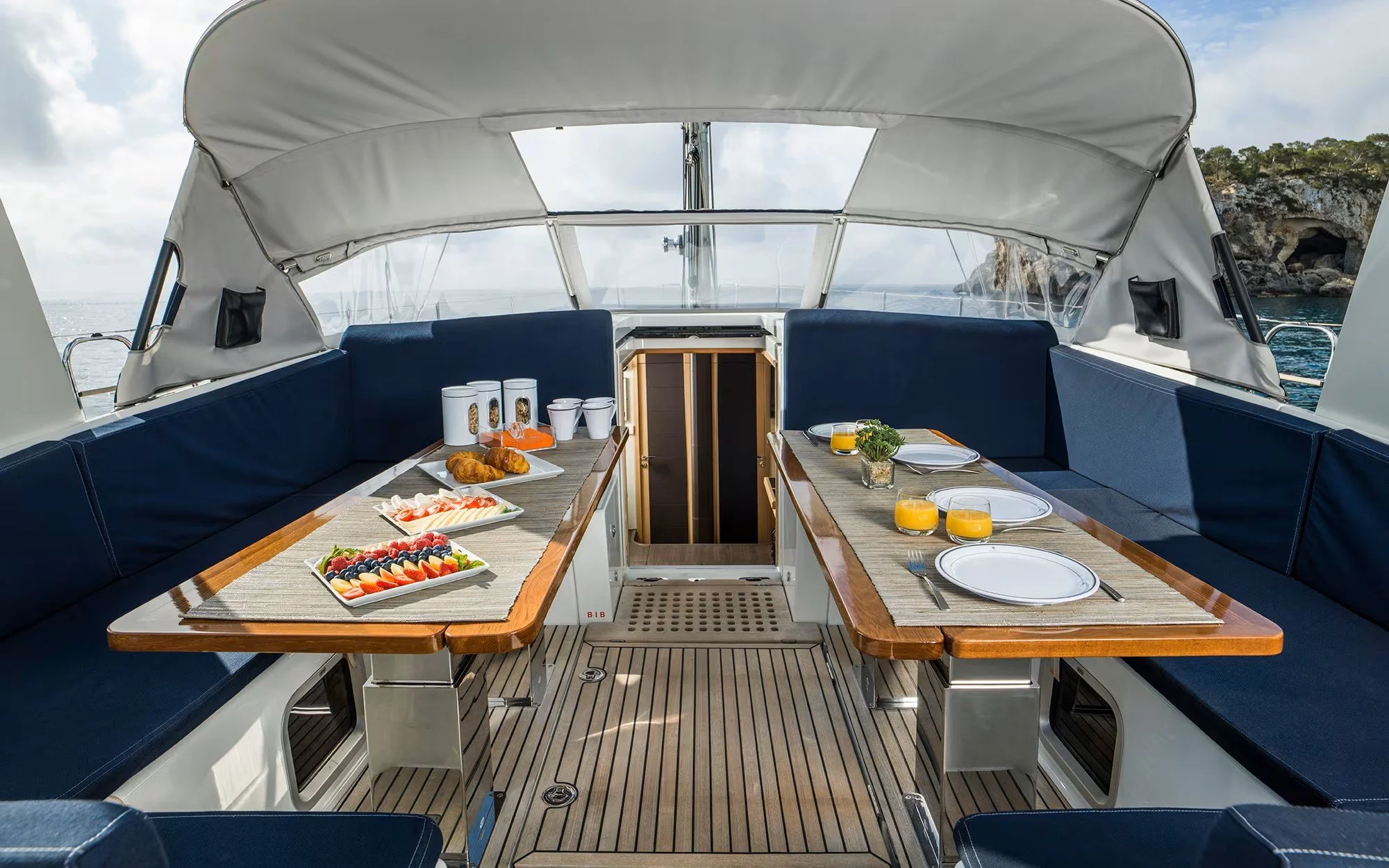 A dining area on a yacht with a wooden table set for breakfast, including plates, glasses of orange juice, and food, surrounded by blue cushioned seating and overlooking the sea.