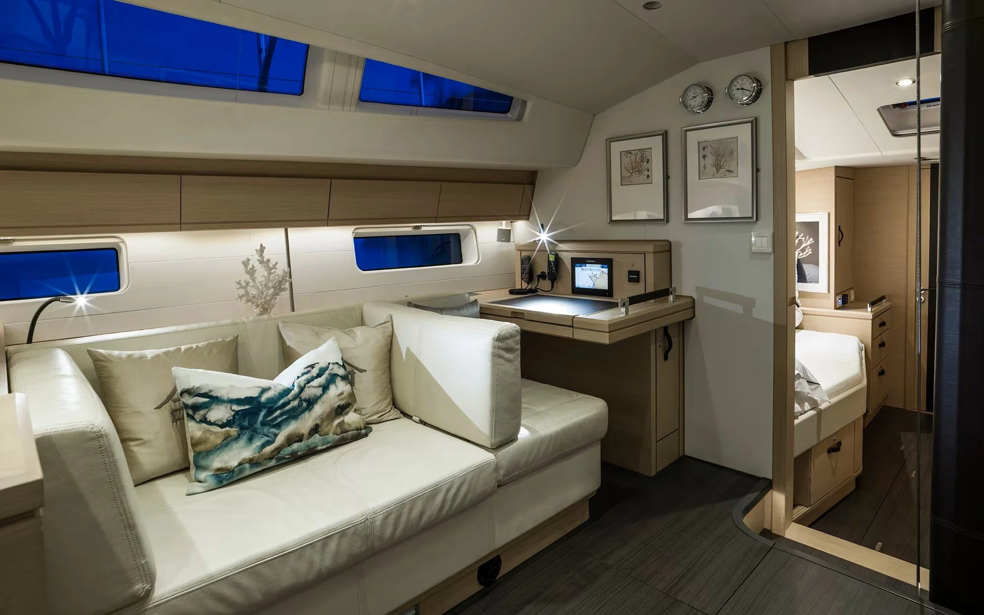 Interior of a modern yacht with a cream sofa, decorative pillows, wooden accents, and a small bedroom visible through an open doorway.