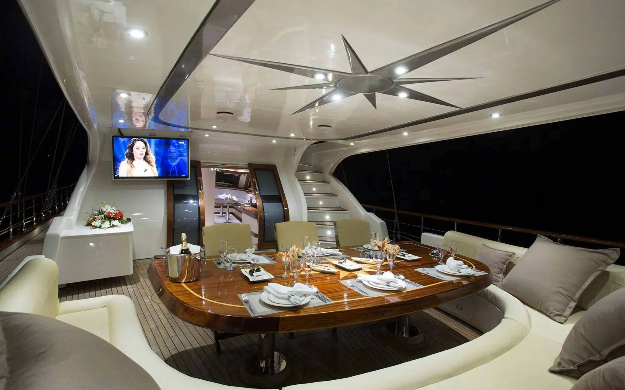 Dining area on a yacht with a large oval wooden table, arranged for a meal with plates, glasses, and napkins, surrounded by cream-colored chairs and sofas, with a flat screen TV and a staircase leading to another deck visible in the background.
