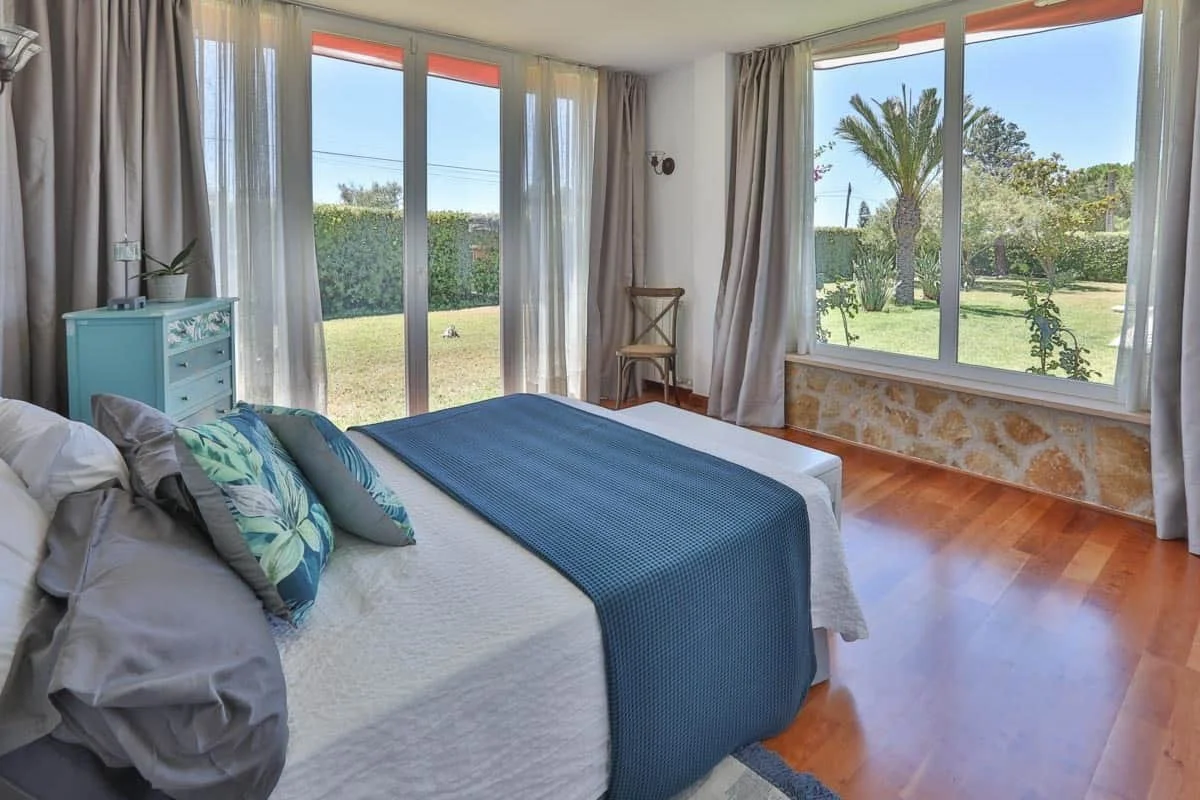 Bright bedroom with which large window offering a view of a lush, green yard including palm trees, shrubs, and a stone wall. The room has wooden floor, light-colored curtains, and a bed with pillows and a blue blanket.
