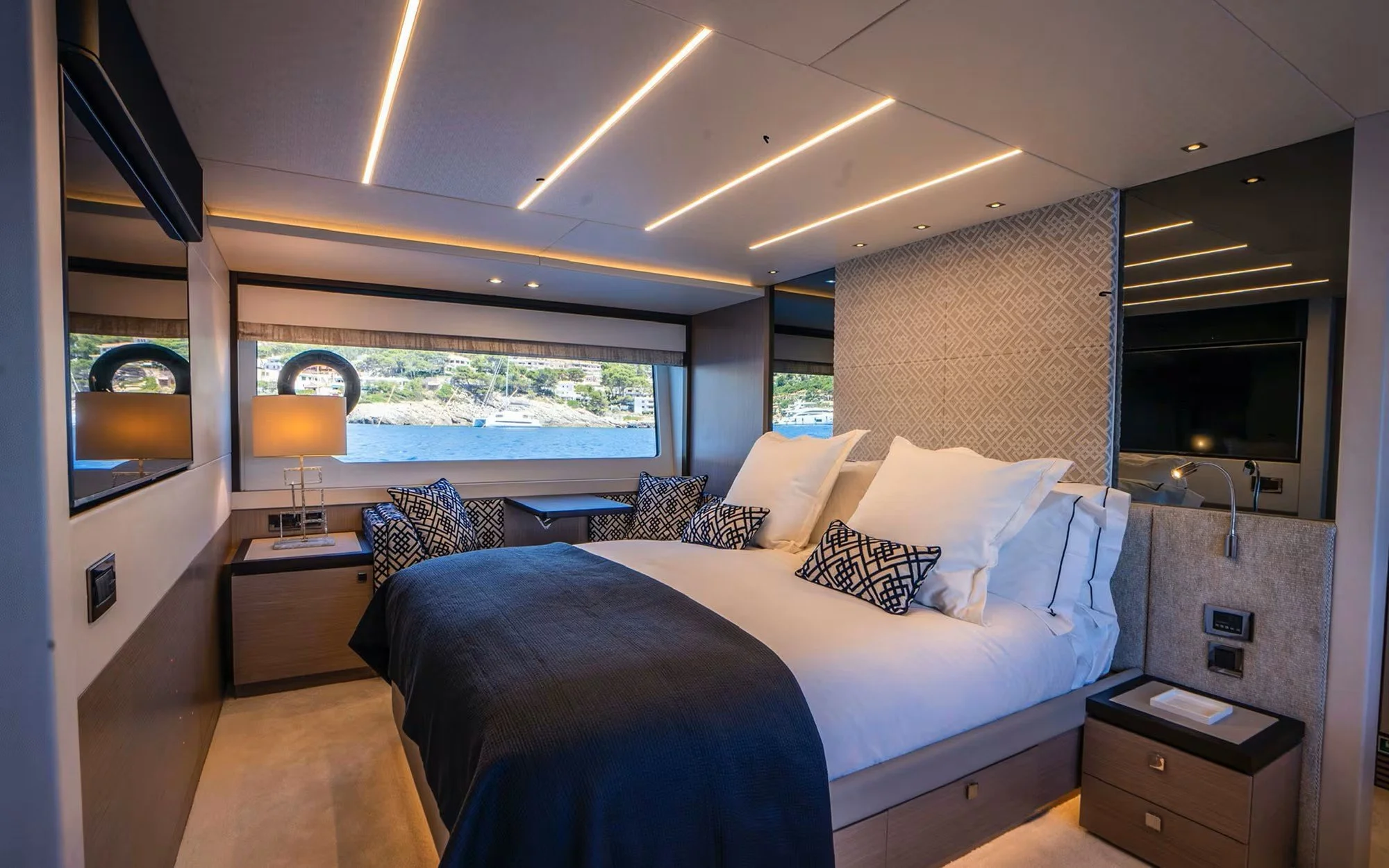 A modern bedroom on a yacht with a large bed dressed in white and dark bedding, decorative pillows, lamps on bedside tables, a wall-mounted flat-screen TV, a panoramic view of water with boats through large windows, and contemporary ceiling lighting.