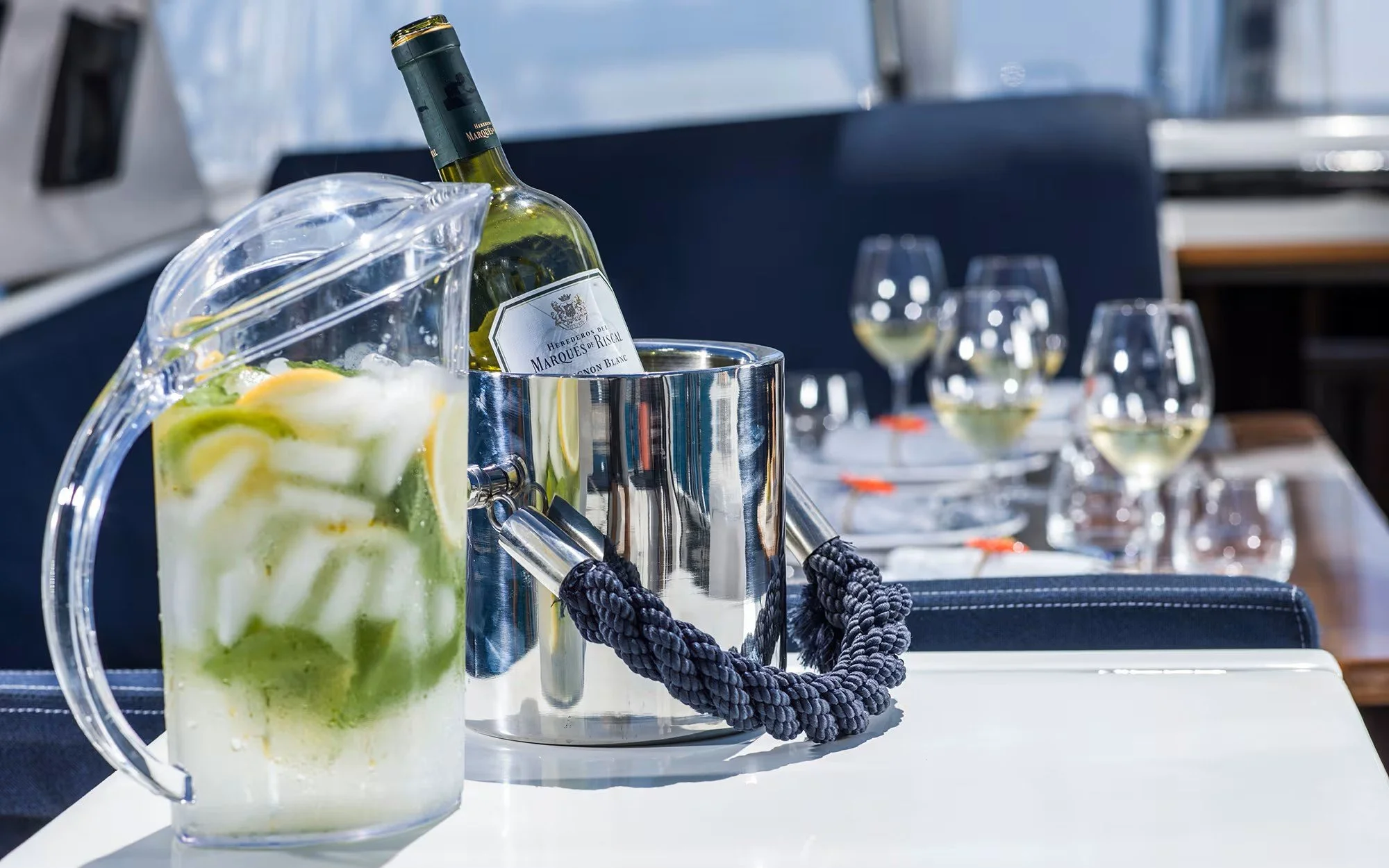 A table with a pitcher of lemon and lime infused water, a bottle of white wine in a metal wine holder, and multiple glasses of white wine.