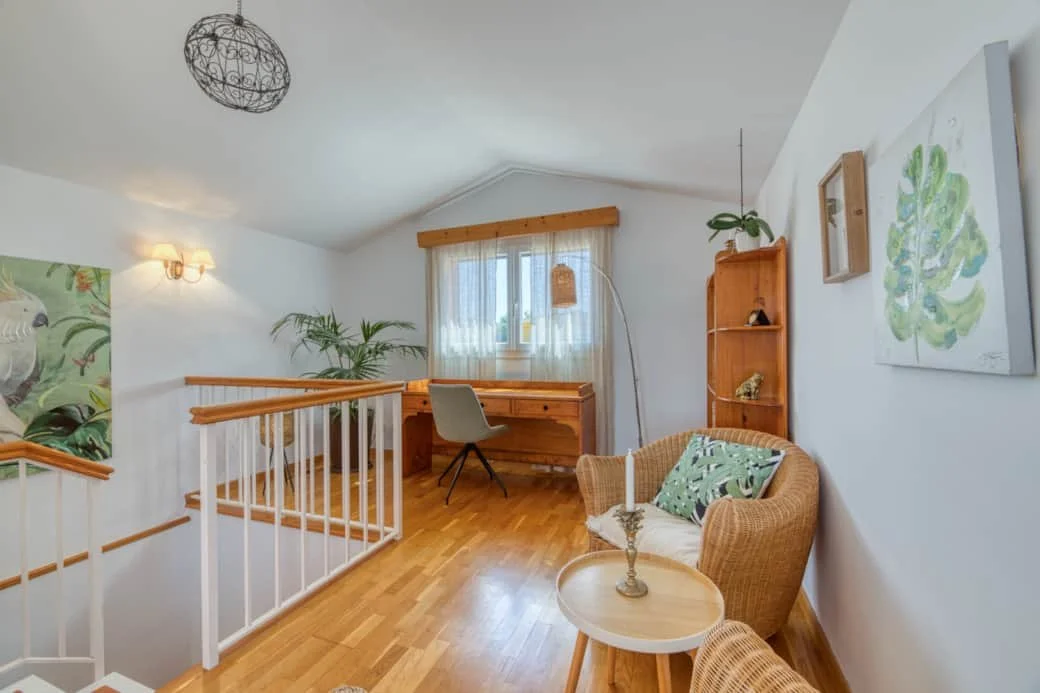 Interior of a home office or reading nook with a wicker chair, a small round wooden table with a candle, a tall bookshelf, a window with sheer curtains, and wooden accents.