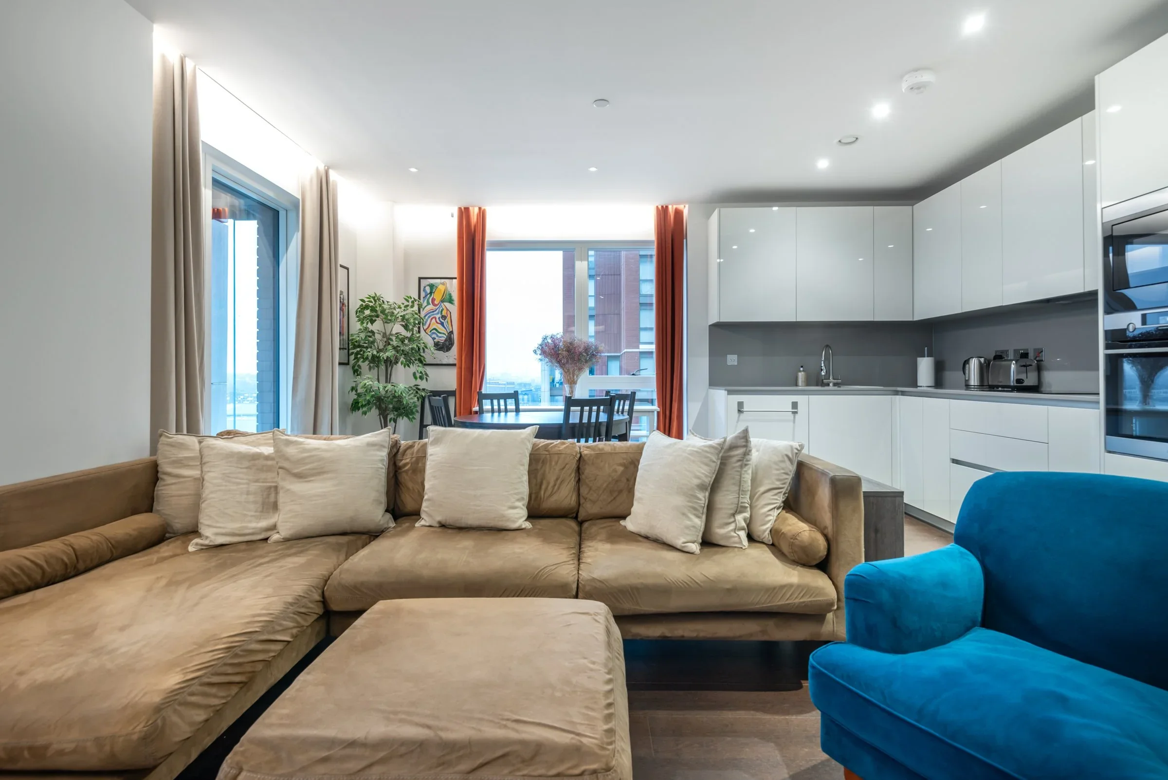 Living room with beige sectional sofa, blue armchair, white cushions, and a view of the kitchen and dining area with a window, plants, and orange curtains.