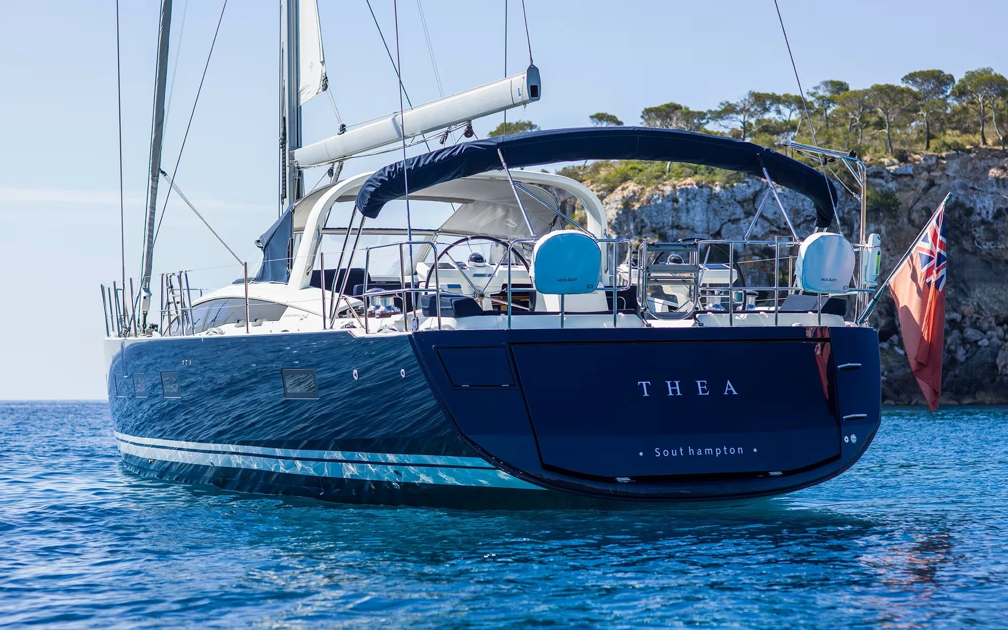 A sailboat with a dark blue hull named THEA, registered in Southampton, sailing near a rocky shoreline with trees on a sunny day.