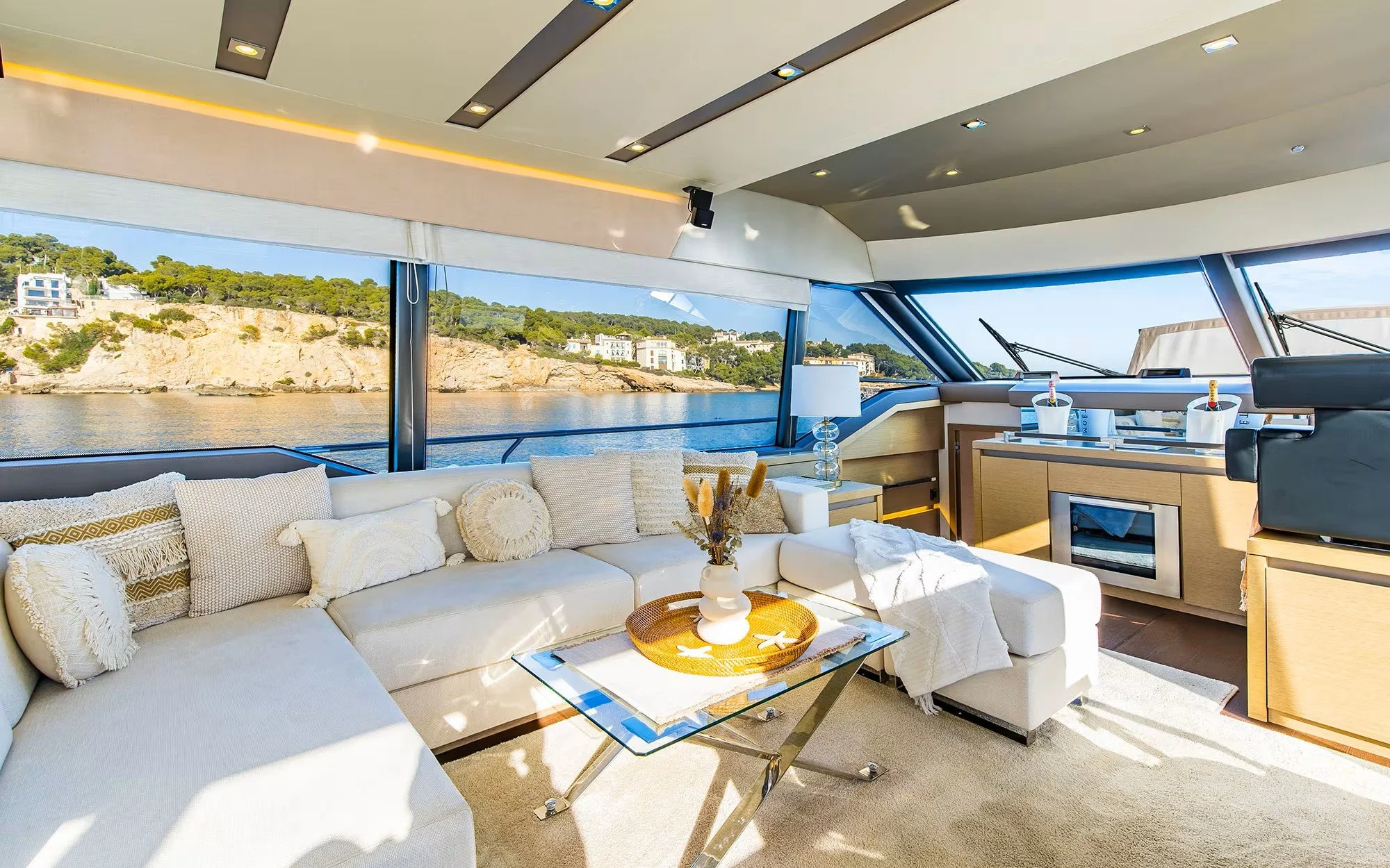 Interior of a yacht with a large white sectional sofa, decorative pillows, a glass coffee table with a flower arrangement, and a view of water and shoreline outside the windows.