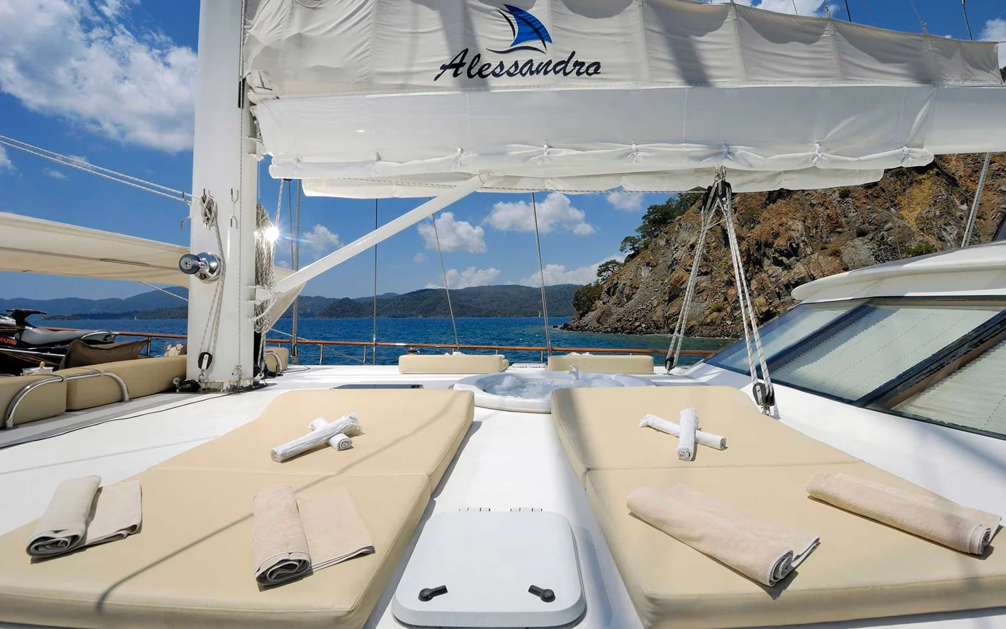 View of a yacht's deck with sunbeds and towels, overlooking blue water and mountainous landscape under partly cloudy sky.