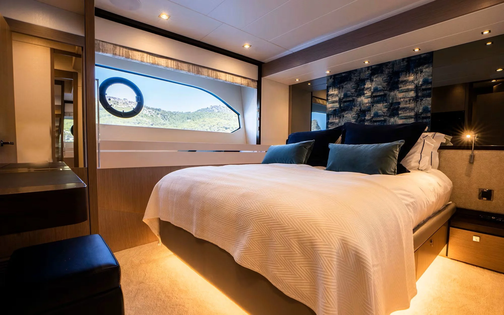 A modern bedroom with a large bed featuring white and dark blue pillows, a wooden headboard, and a window with a scenic view of mountains and hills.