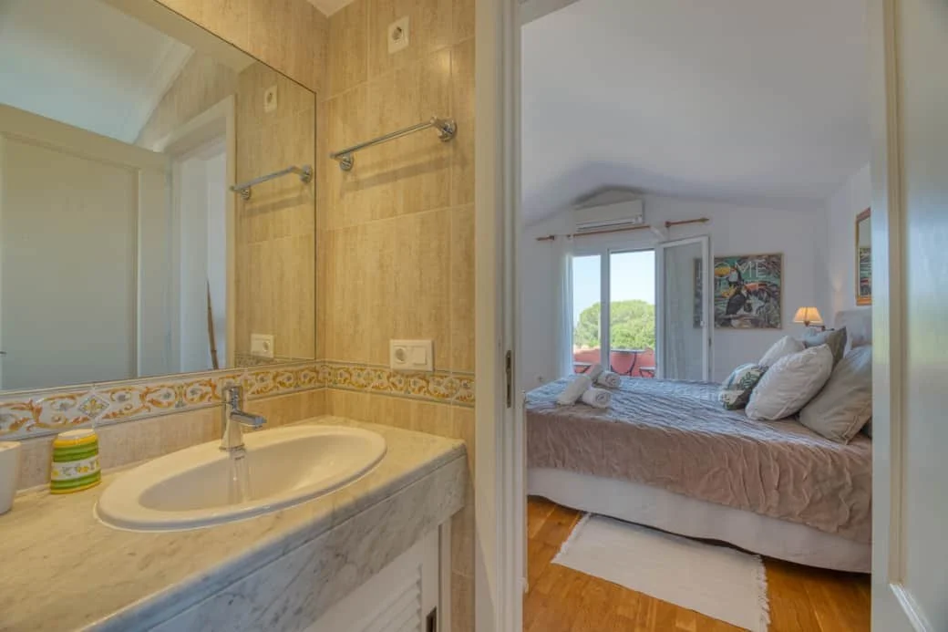 Bathroom with a marble countertop, sink, towel rack, and mirror, adjacent to a bedroom with a bed, window with curtains, and wall art.