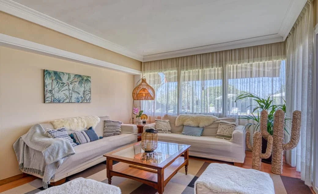 Living room with two white sofas, throw blankets and multiple pillows, a wooden coffee table, large windows with sheer curtains, a cactus sculpture, and tropical artwork on the wall.