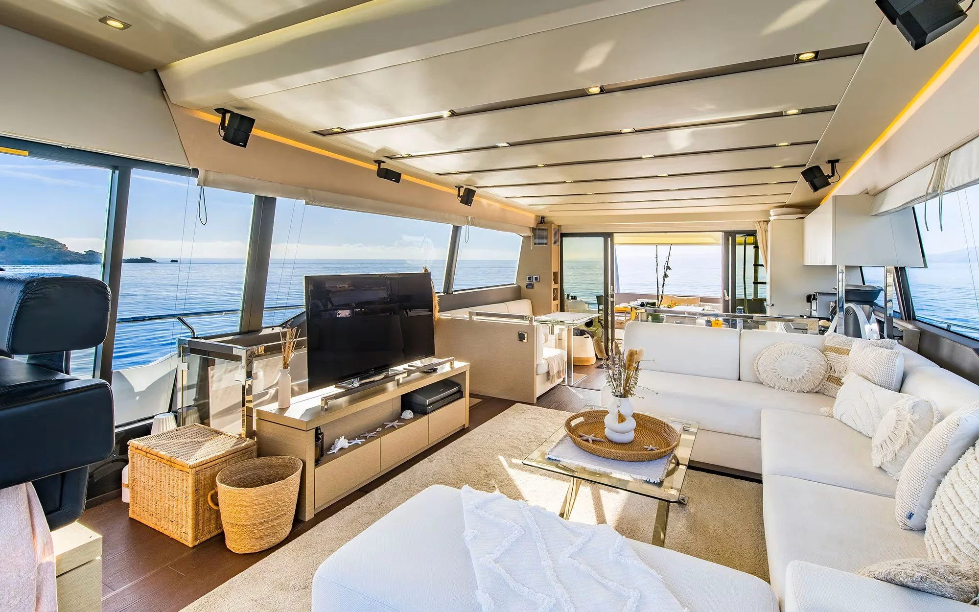 Bright living room on a boat with large windows showing the ocean outside, a white sectional sofa with decorative pillows, a glass coffee table, and a flat-screen TV on a stand.