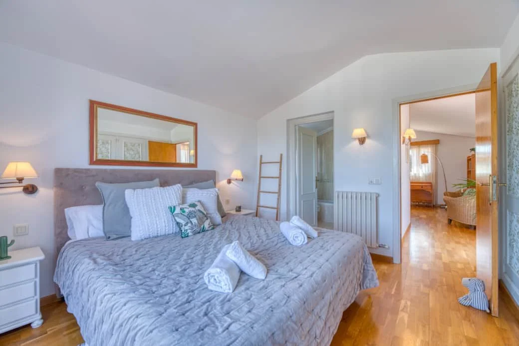 A cozy bedroom with a large bed dressed in gray bedding, white and patterned pillows, and neatly folded towels. The room has white walls, wooden accents, a mirror above the headboard, and a wooden ladder leaning against the wall. There's a small whit