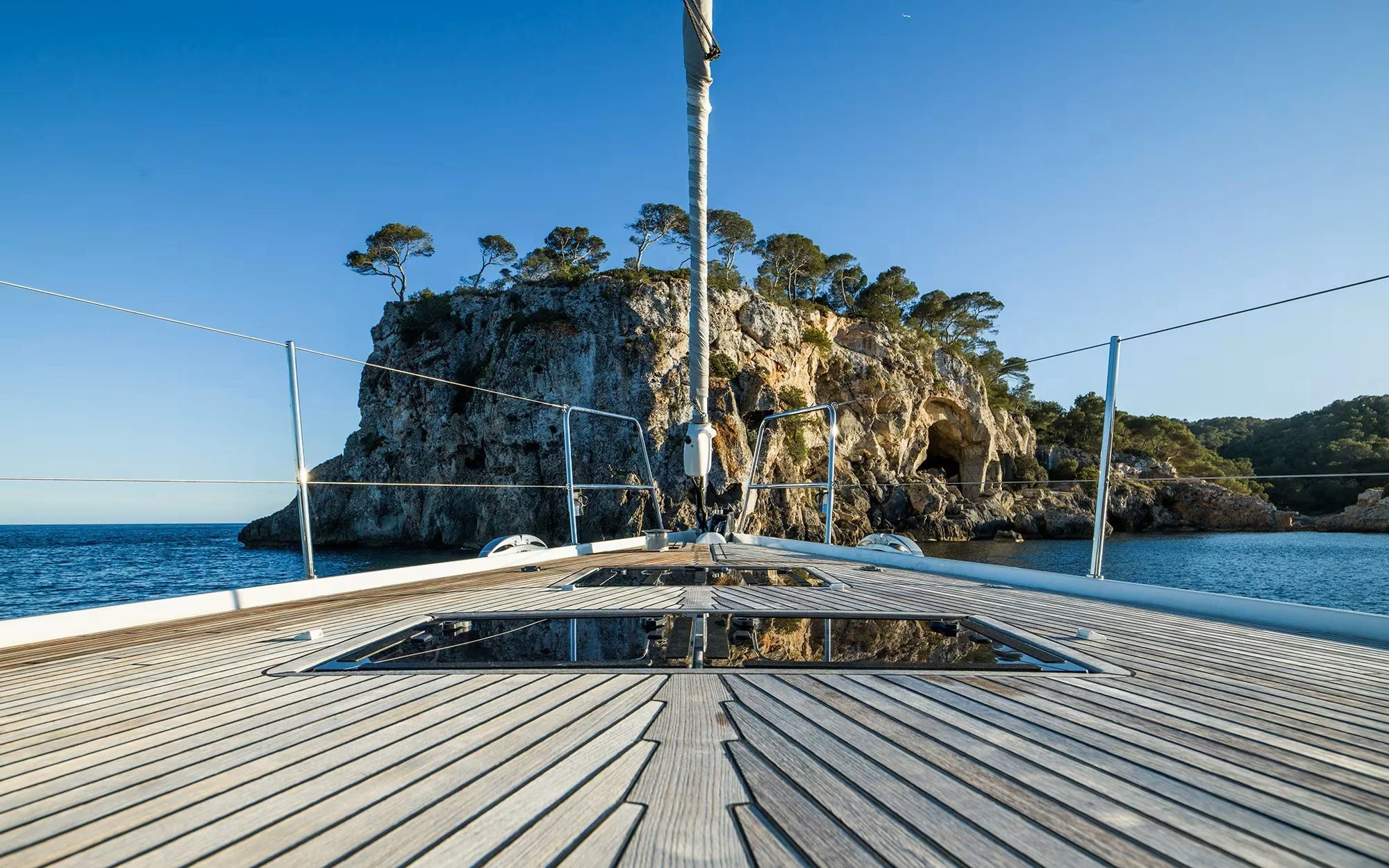 View from the deck of a sailing yacht showing the wooden surface, with a rocky island with trees in the background, calm water, and a clear blue sky.