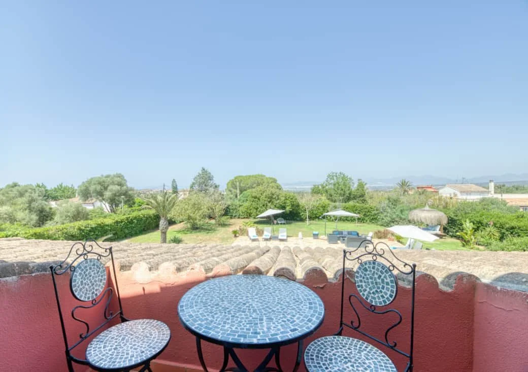View of a garden with trees, umbrellas, and lounge chairs seen from a balcony with a red wall, featuring a small round mosaic table and two mosaic chairs.