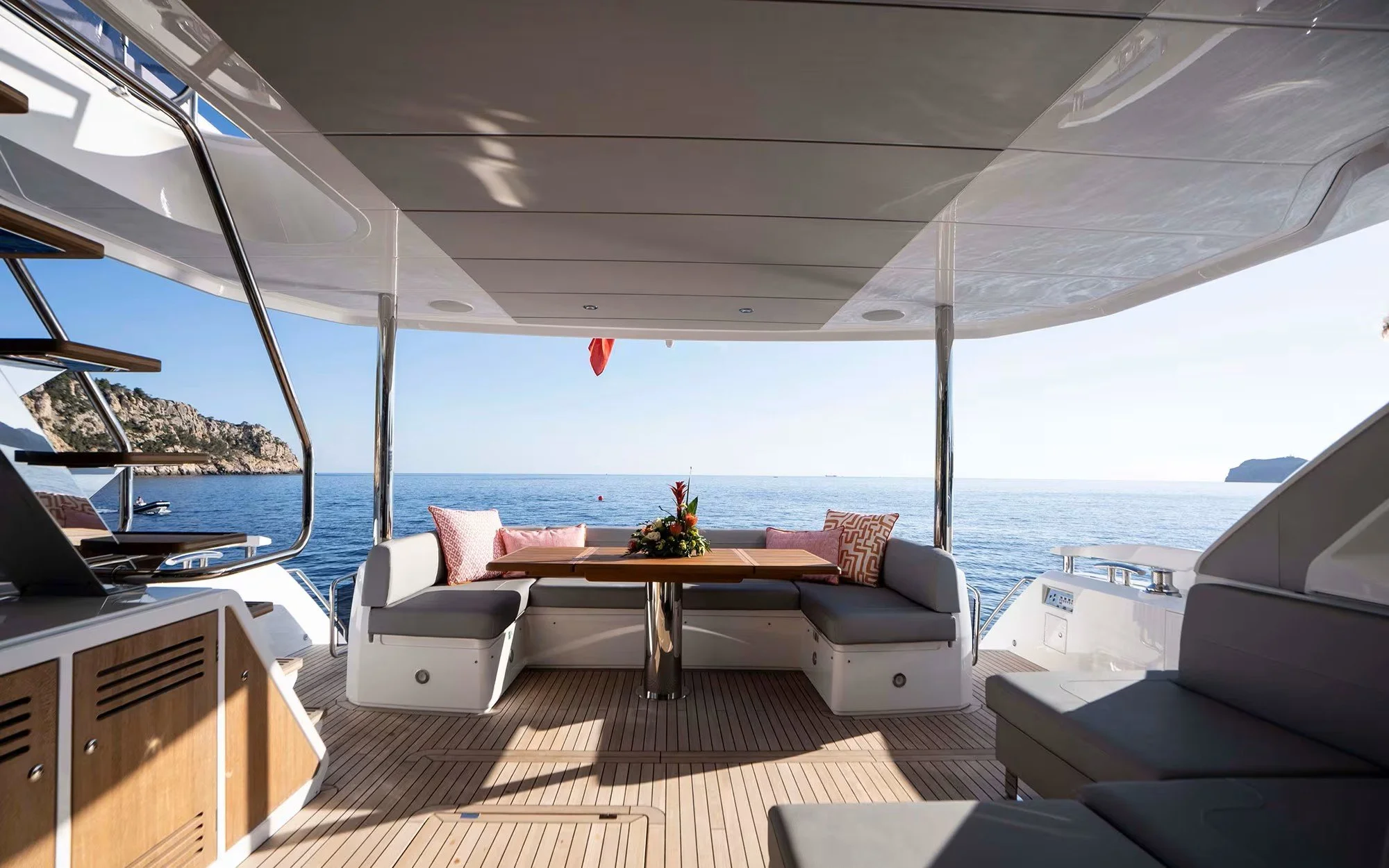 Outdoor seating area on a yacht with a table, decorative pillows, and a view of the ocean and distant land.