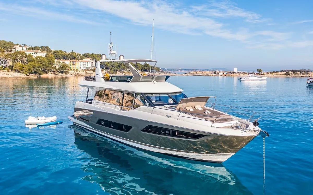 A large white yacht anchored near a shoreline with houses, with several smaller boats floating nearby on calm blue water under a partly cloudy sky.