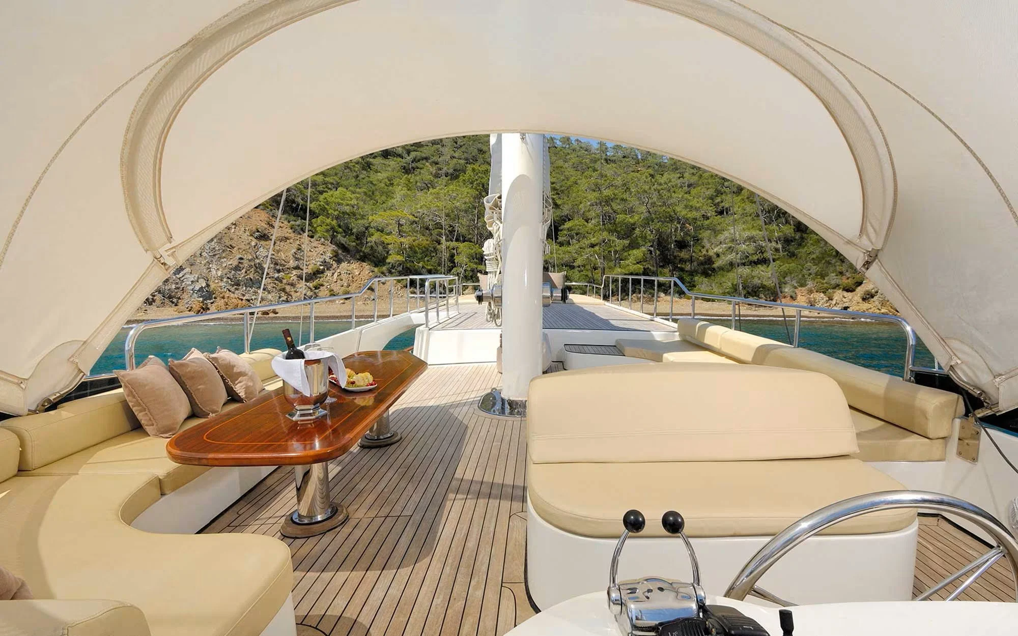 View from on deck of a yacht with beige cushioned seating, a wooden table with a wine bottle and tray of food, overlooking trees and water