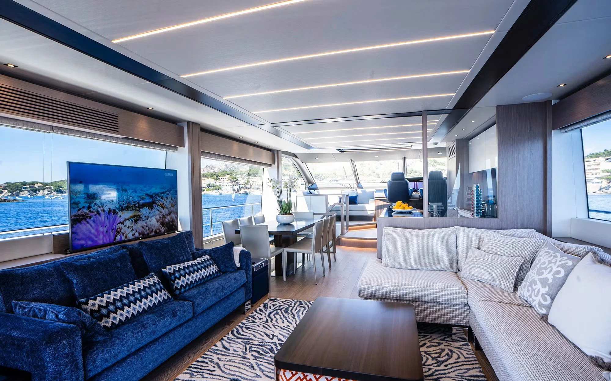 Interior of a modern yacht with a view of the water and shoreline outside. Furnished with a blue velvet sofa, a beige sectional sofa, a wooden coffee table, and a dining table with chairs. The ceiling has integrated lighting, and large windows let in
