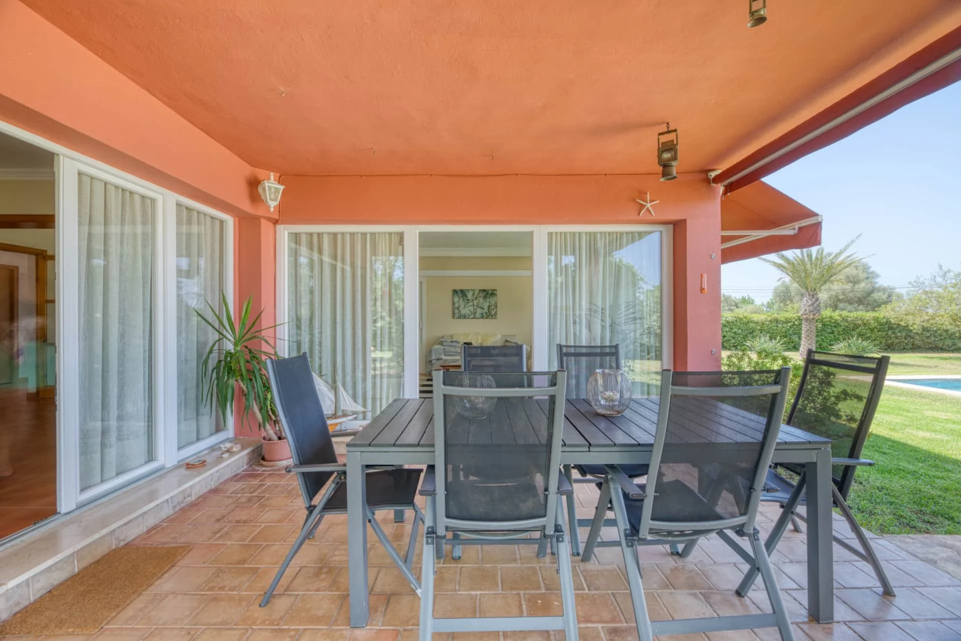 Covered patio with outdoor dining table and chairs, potted plant, sliding glass doors, view of backyard with palm tree and swimming pool.