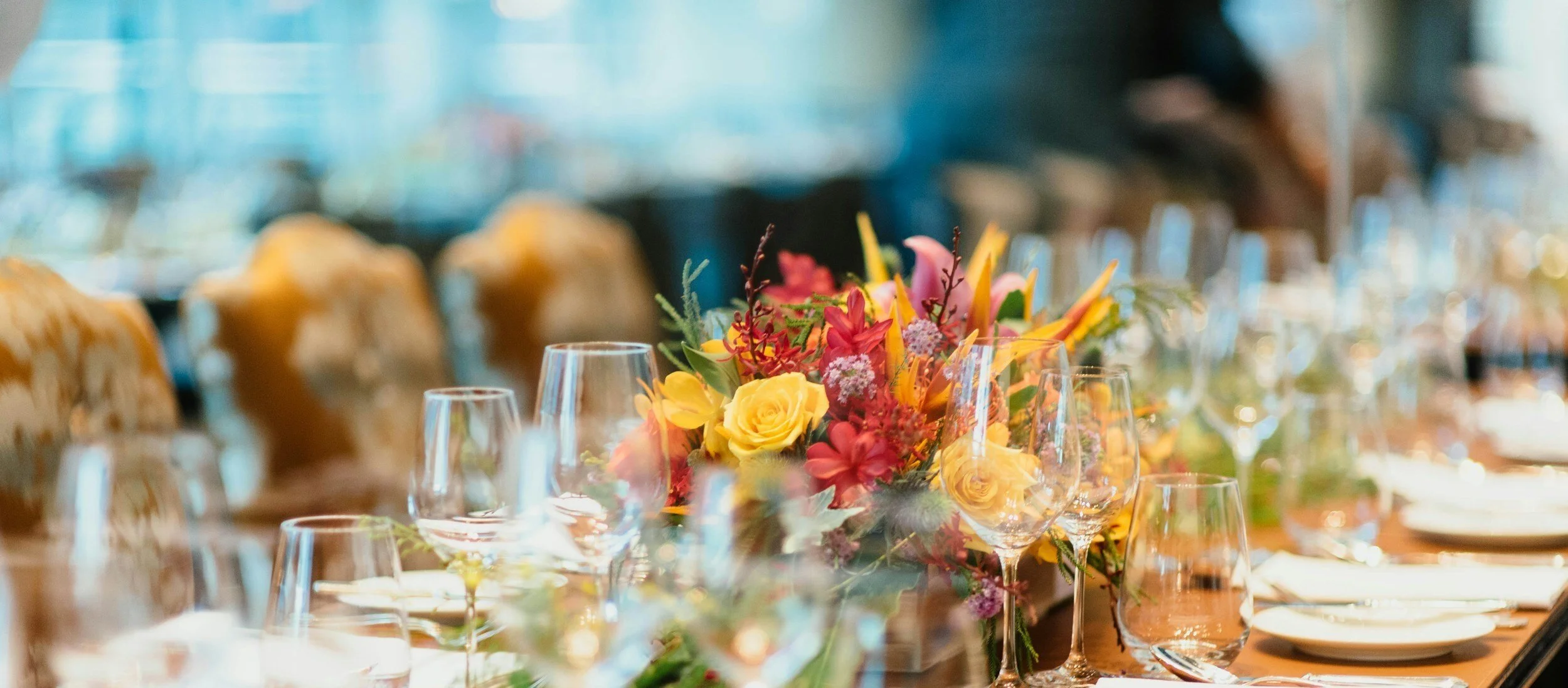 A banquet table set with empty wine glasses, white plates, and a colorful floral centerpiece, with blurred chairs and background.
