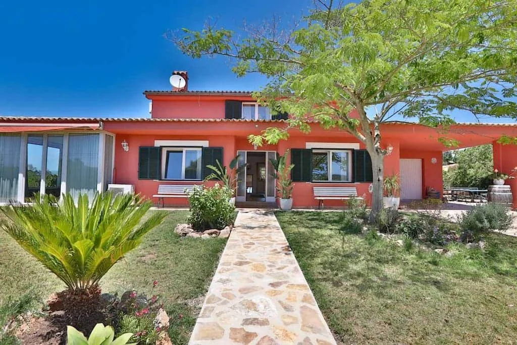 A bright pink house with black shutters, a stone pathway leading to the front door, and a well-maintained lawn with various plants and trees under a clear blue sky.