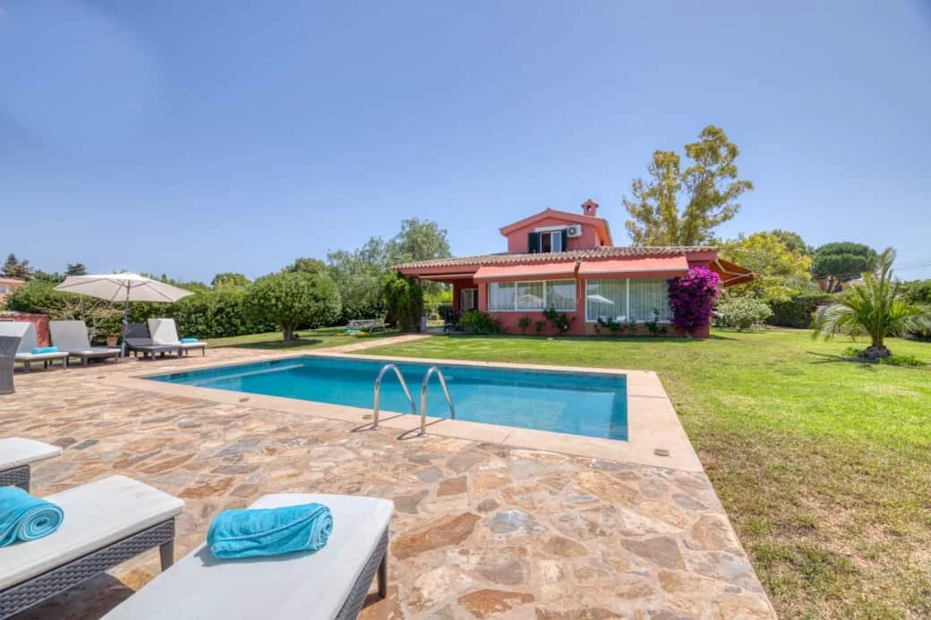 A backyard with a swimming pool and lounge chairs, a pink house with a red roof, surrounded by green trees and bushes, under a clear blue sky.